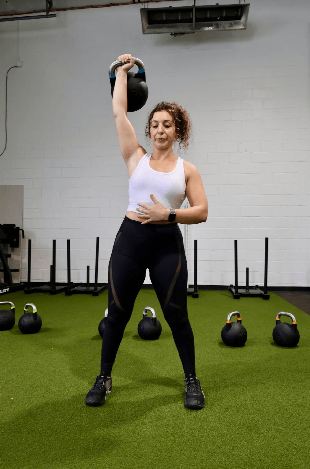 Woman lifting a kettlebell overhead in a gym with kettlebells on green floor behind her.