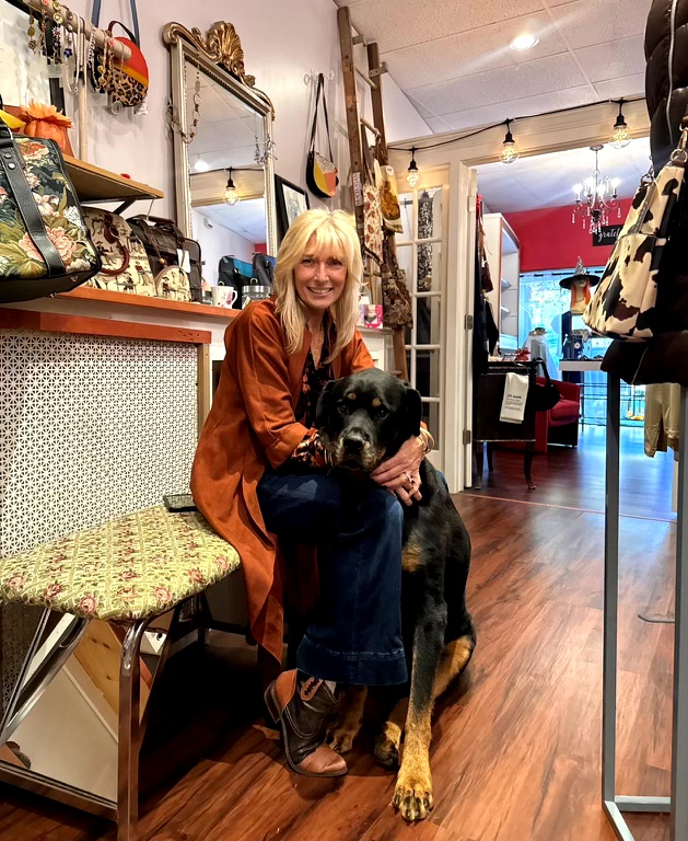 Woman sitting on a bench with a large black and tan dog inside a boutique store, surrounded by clothing and accessories.
