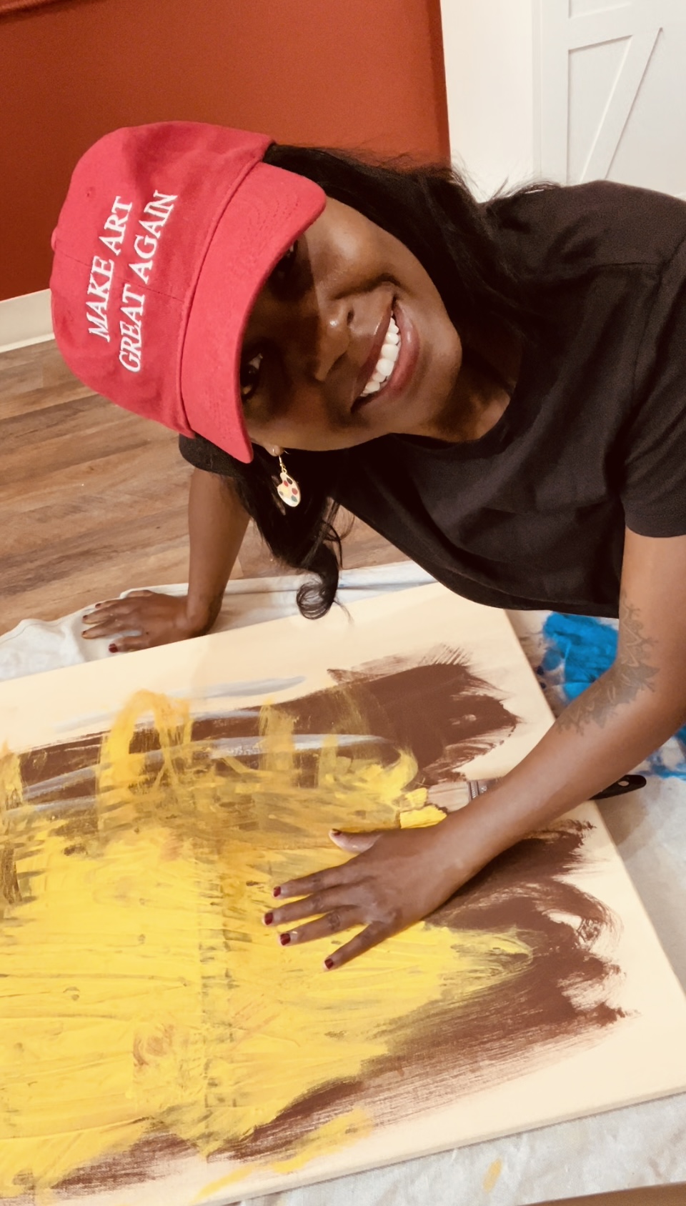 Person smiling, wearing a red cap and black shirt, painting with yellow and brown colors on canvas.