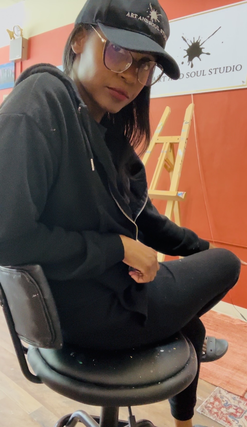 Person wearing glasses, cap, and hoodie sitting on a chair indoors, with a ladder and sign in background.