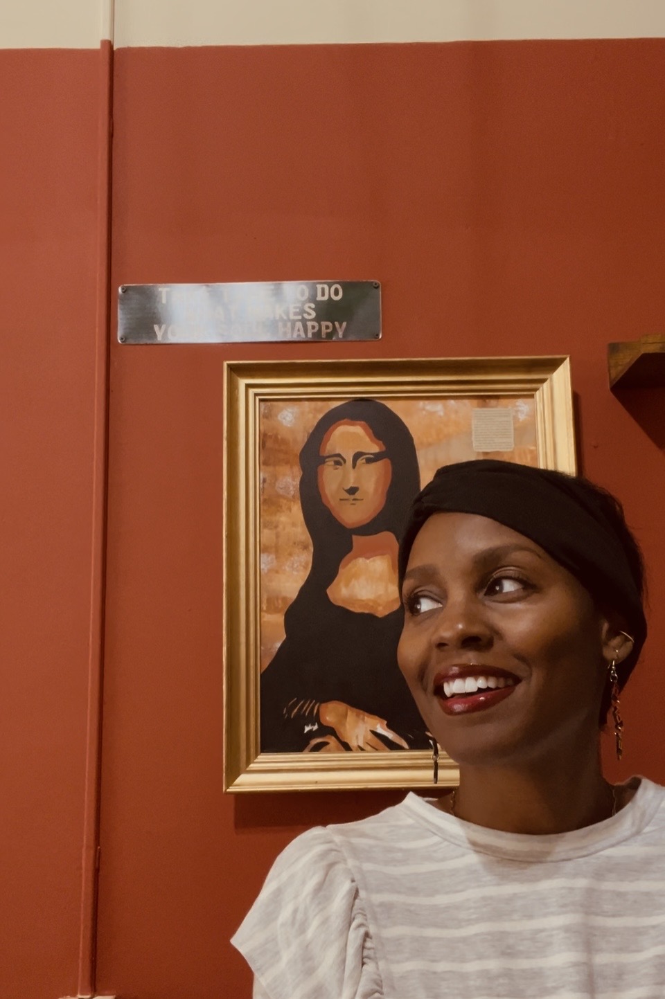 A woman with a headscarf smiling in front of a framed painting of Mona Lisa, against a red wall.