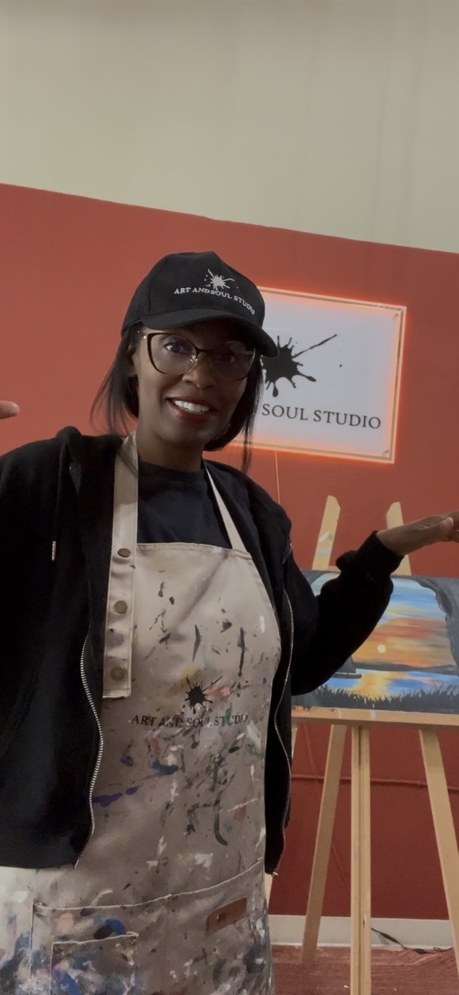 Person wearing glasses, a black cap, and apron, standing indoors with a painting on an easel behind them.