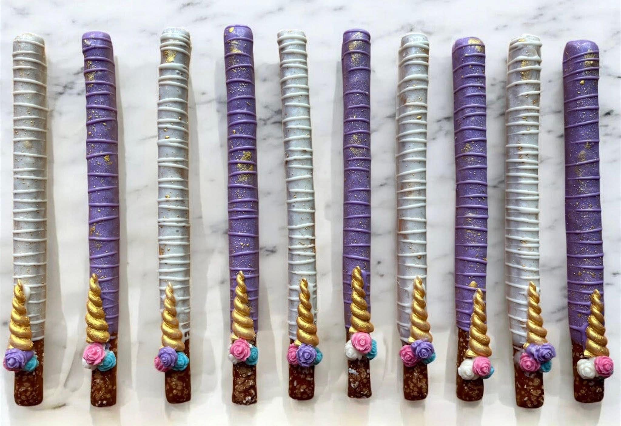 Rows of decorated cake pops with purple, white, and gold colors, arranged on a marble surface.