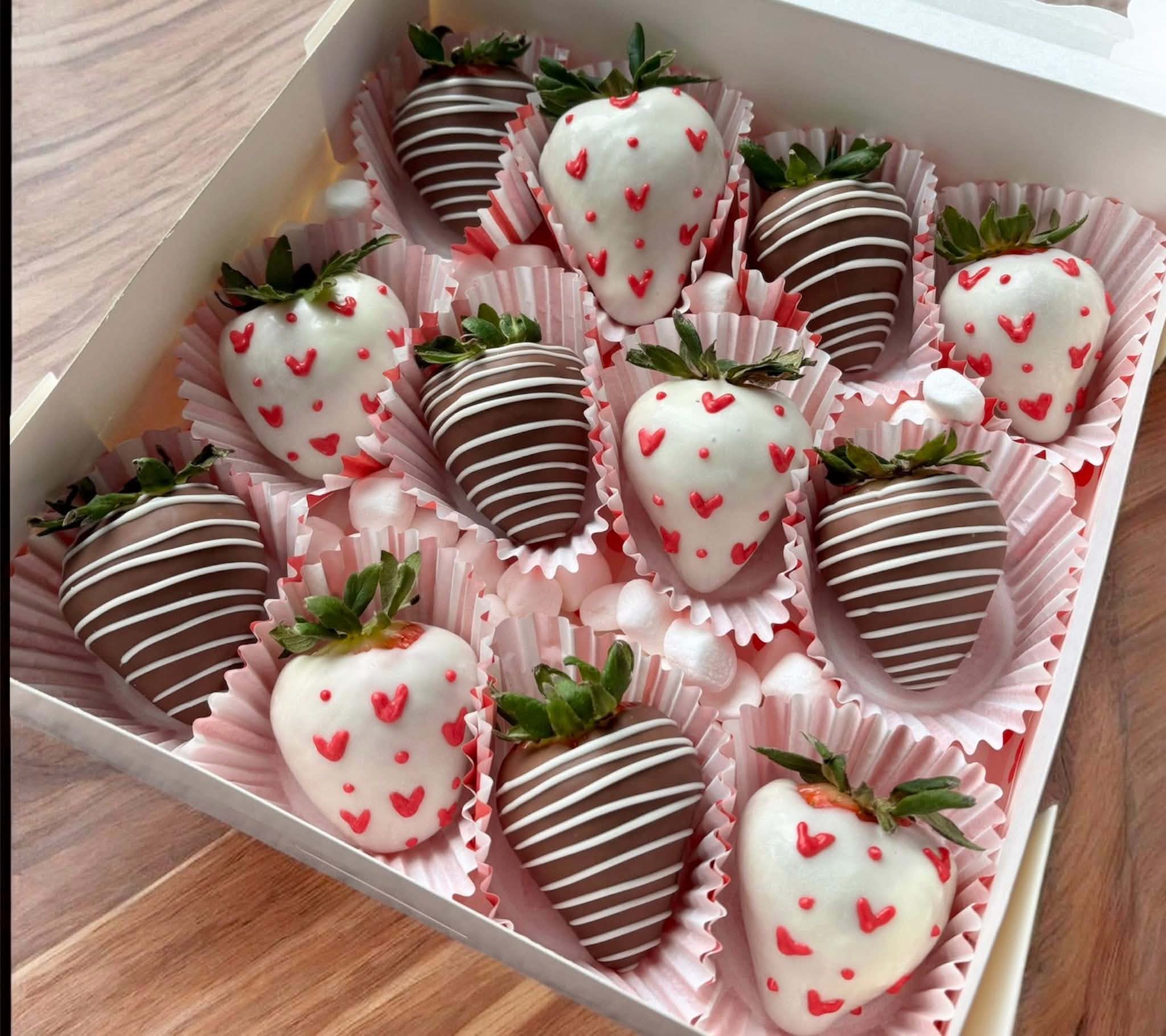 Box of strawberries dipped in chocolate, decorated with white and red hearts, arranged in pink paper cups.