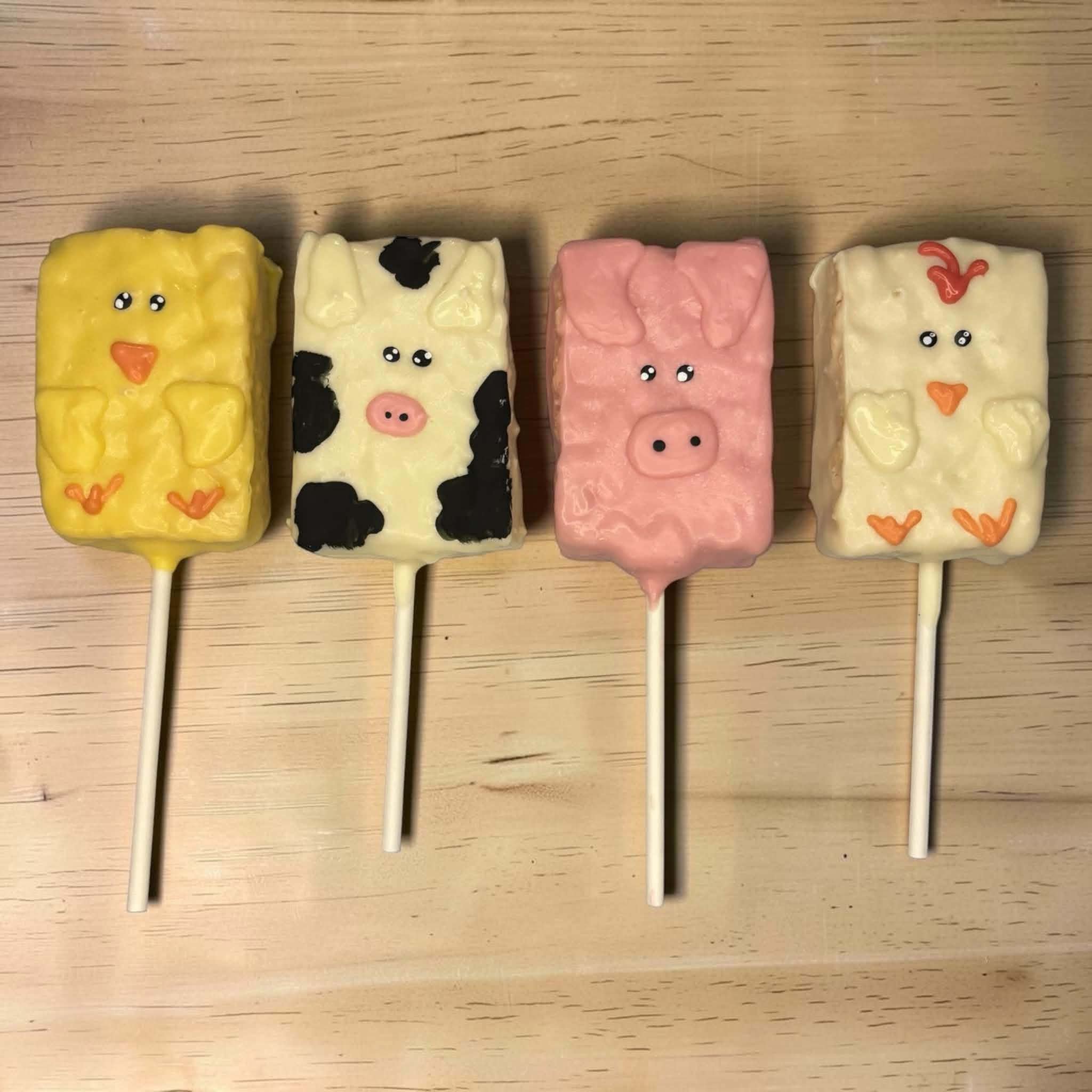 Four animal-shaped marshmallow pops on sticks, resembling a yellow bear, cow, pink pig, and white rabbit, on a wooden surface.