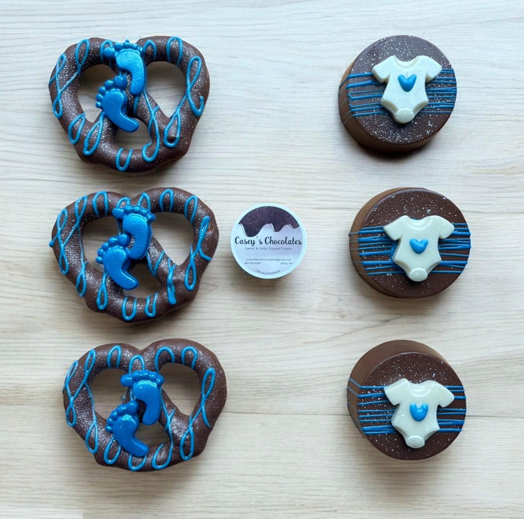 Three blue and brown heart-shaped cookies with blue icing, three round chocolate cookies with white baby onesie decorations, and a small round label in the center.