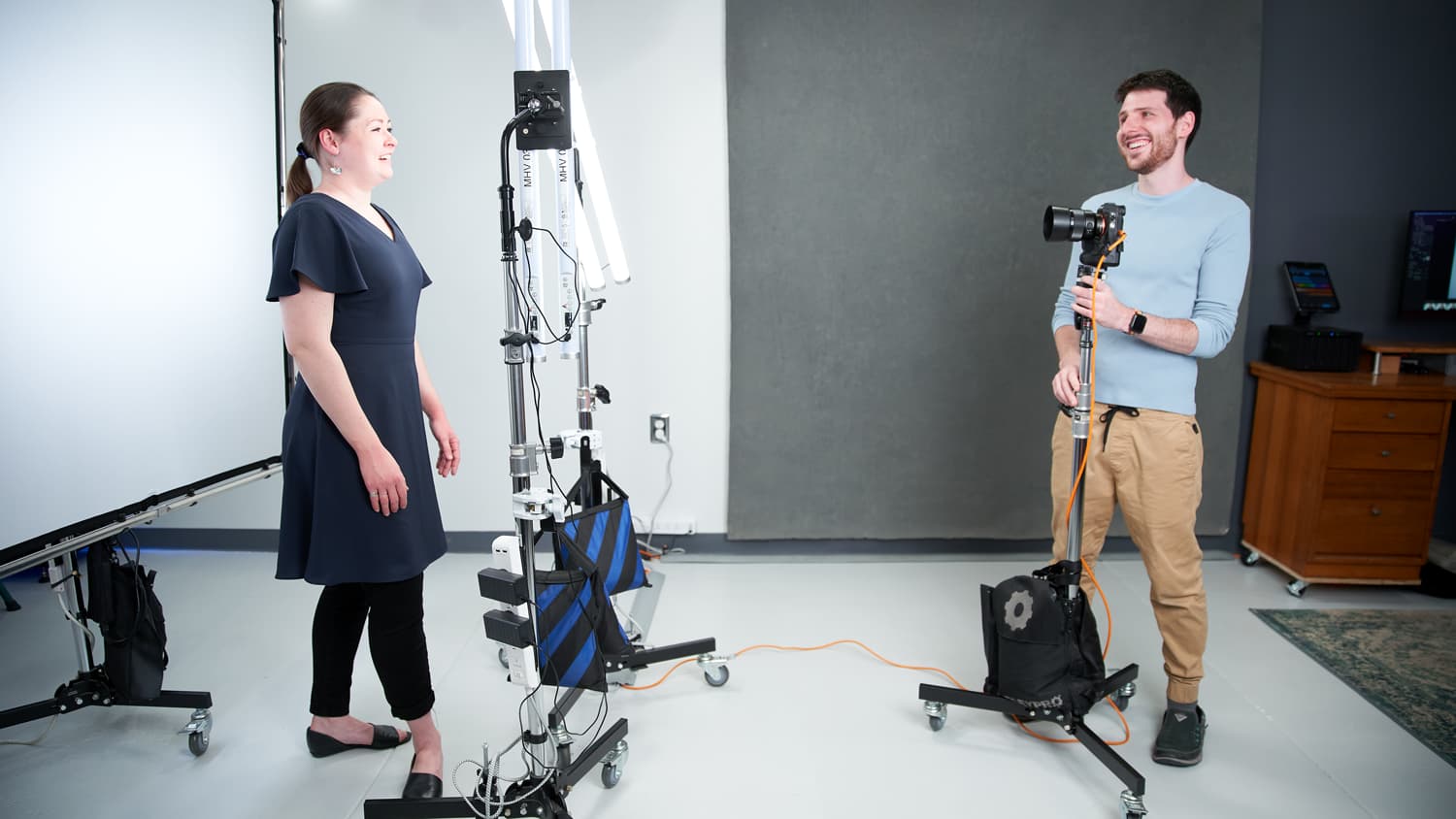 A woman in a dark dress faces a man holding a camera in a studio setting.
