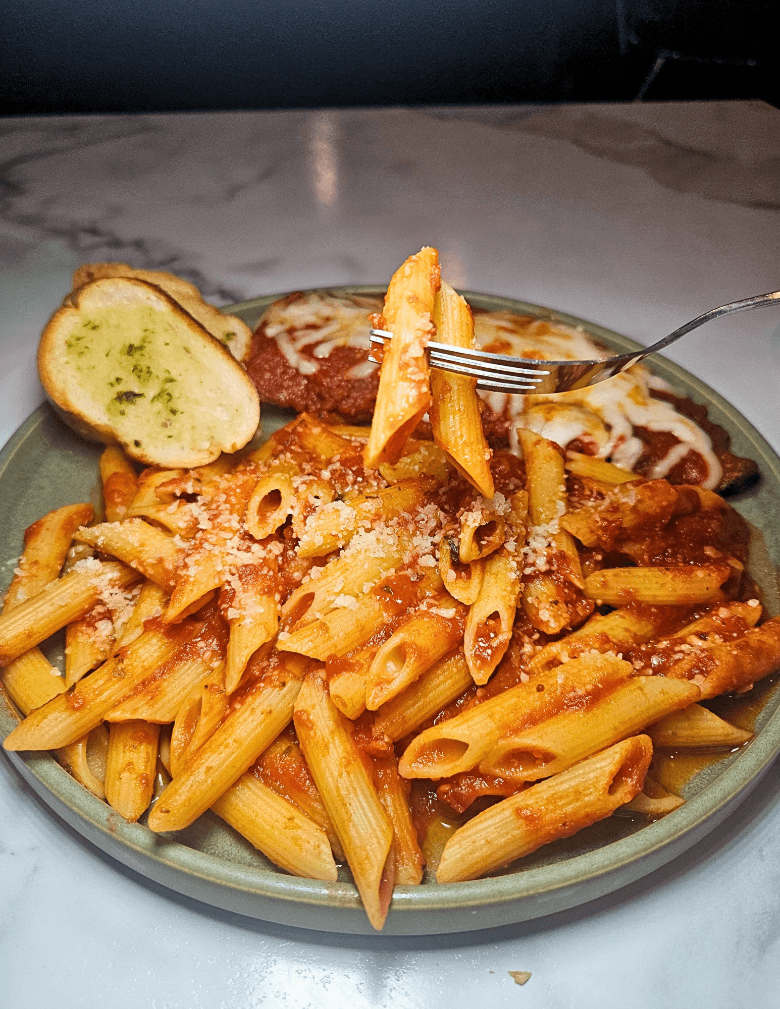 Plate of cooked penne pasta with tomato sauce, grated cheese, a slice of garlic bread, and a fork holding pasta.