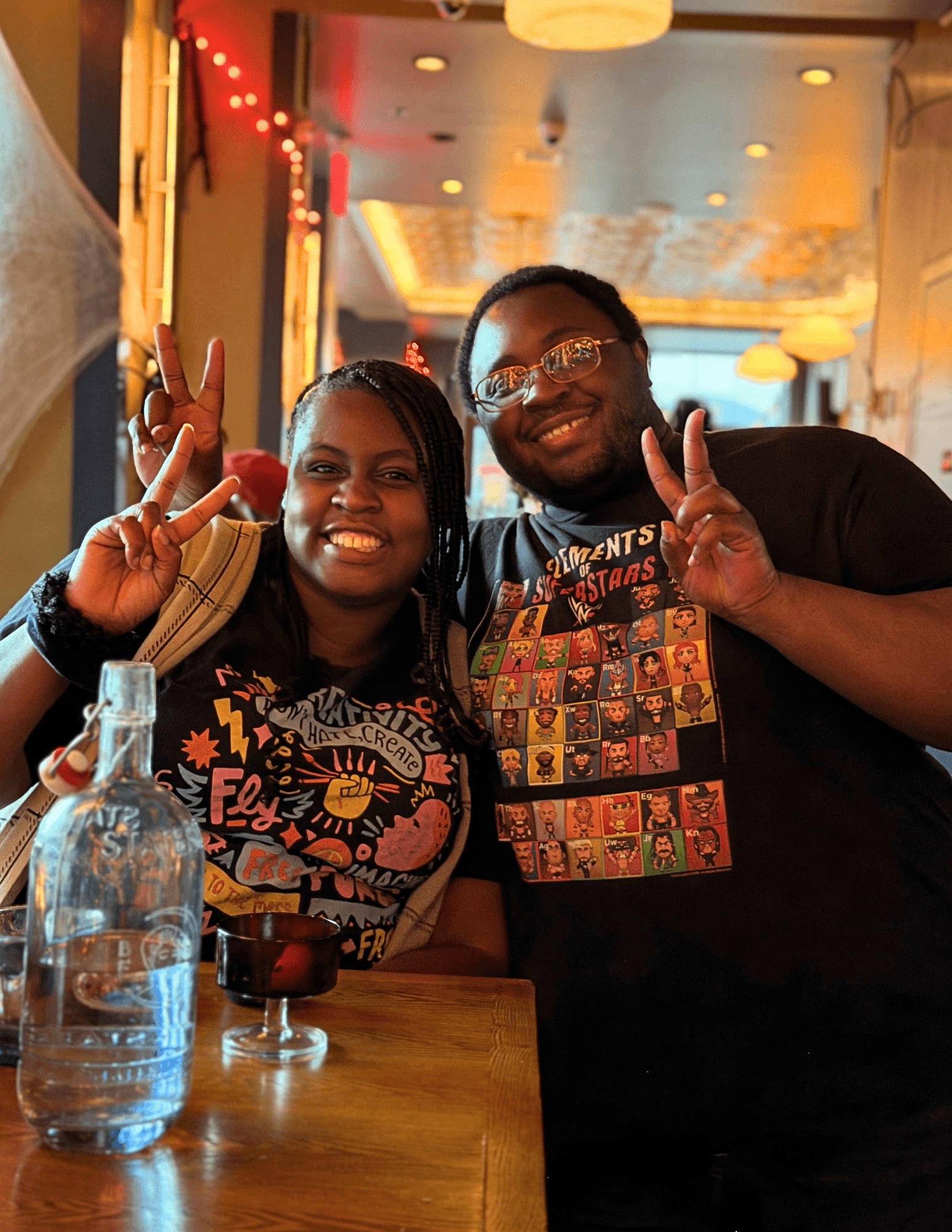 Two smiling people making peace signs in a restaurant, with a table, water bottle, and colorful background.