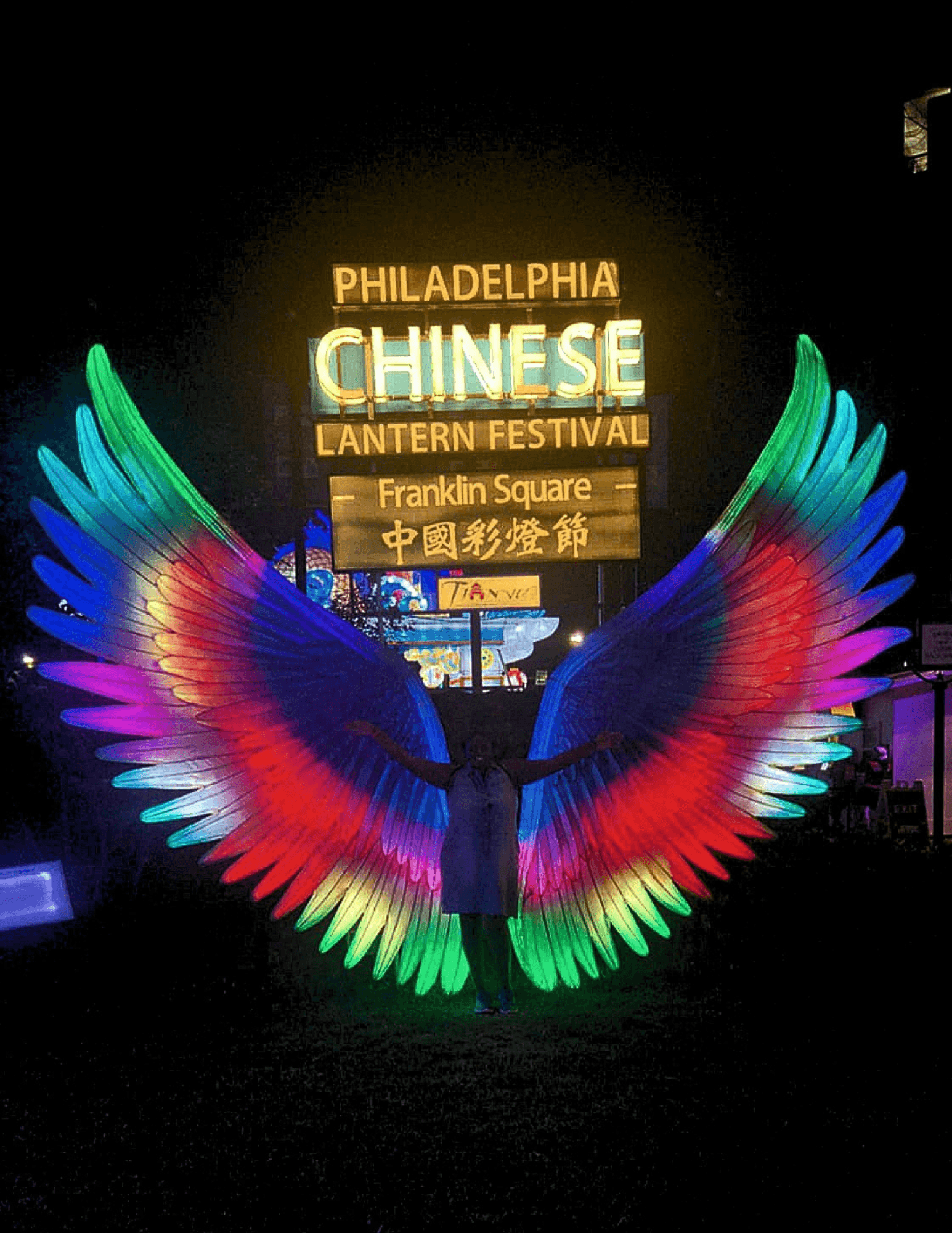 Person standing with illuminated, colorful LED wings at night during Philadelphia Chinese Lantern Festival.