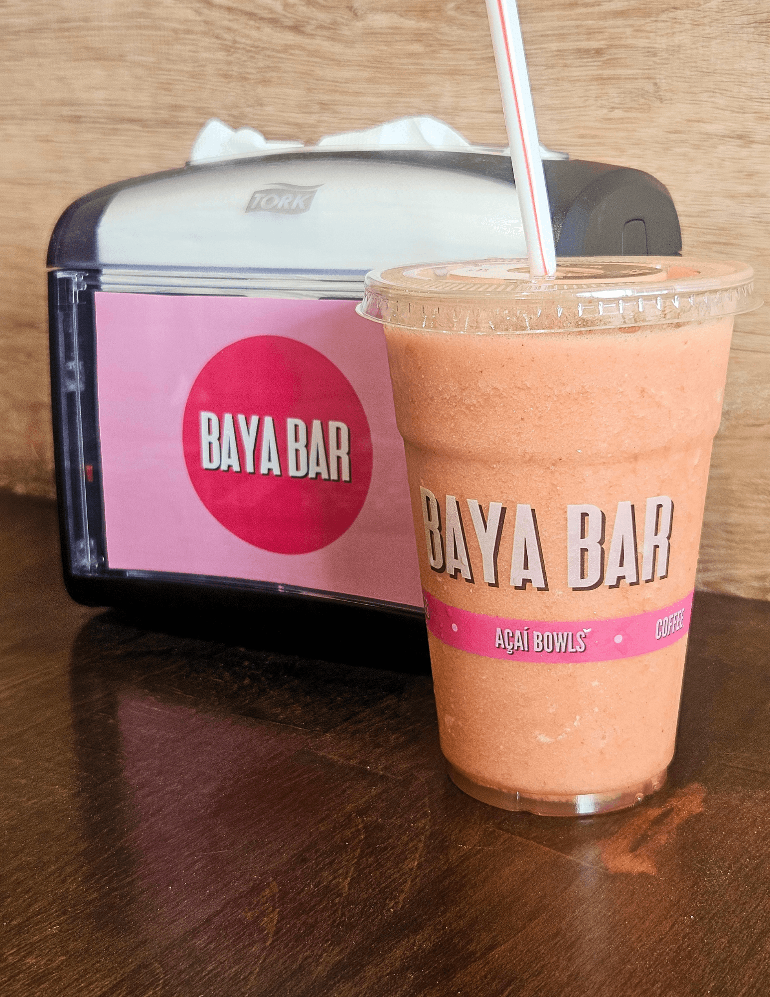 A pink and orange smoothie in a clear cup with a straw, placed on a wooden surface, with a pink Baya Bar container in the background.