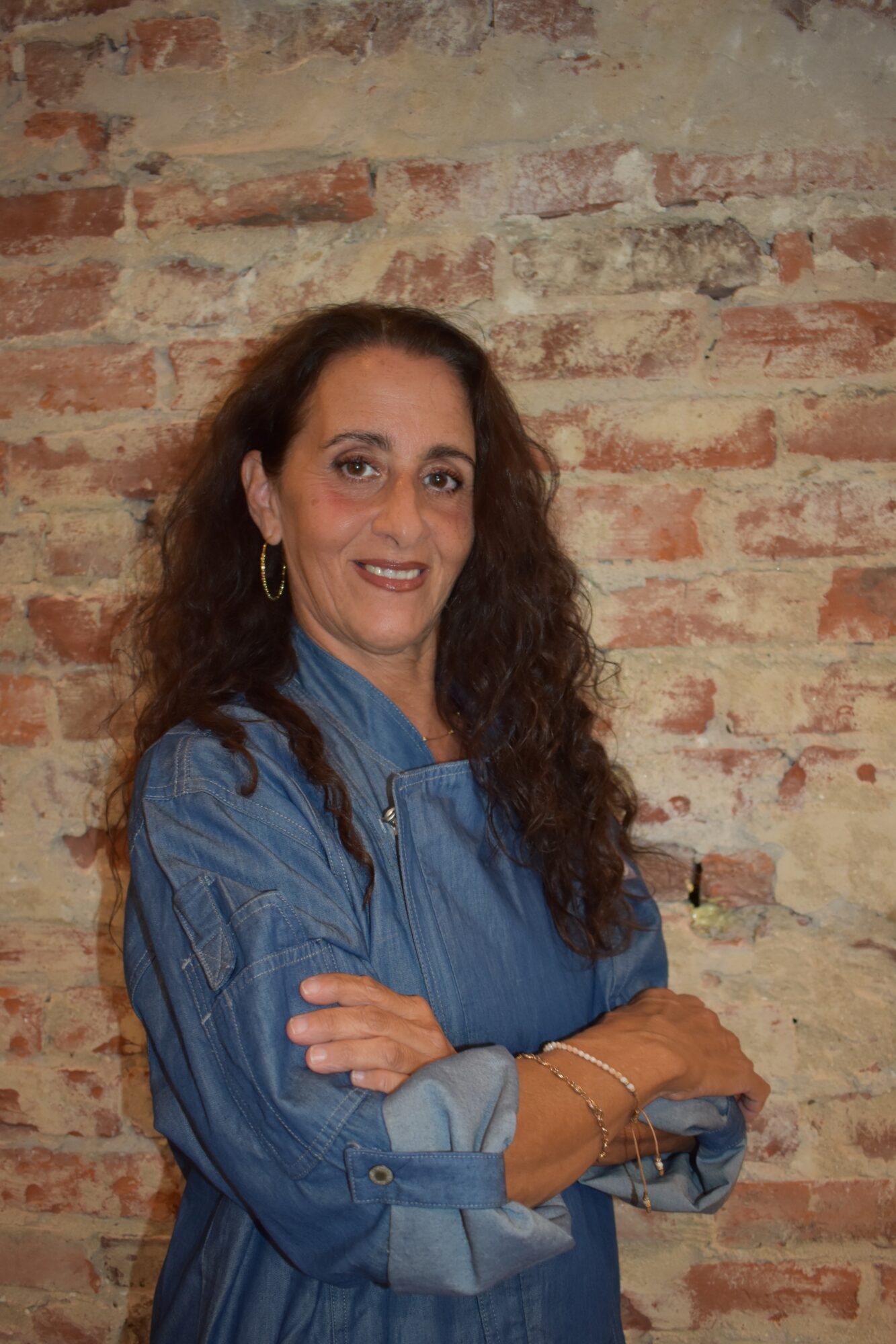 Woman with curly hair wearing a blue denim shirt, standing against a brick wall, smiling with arms crossed.