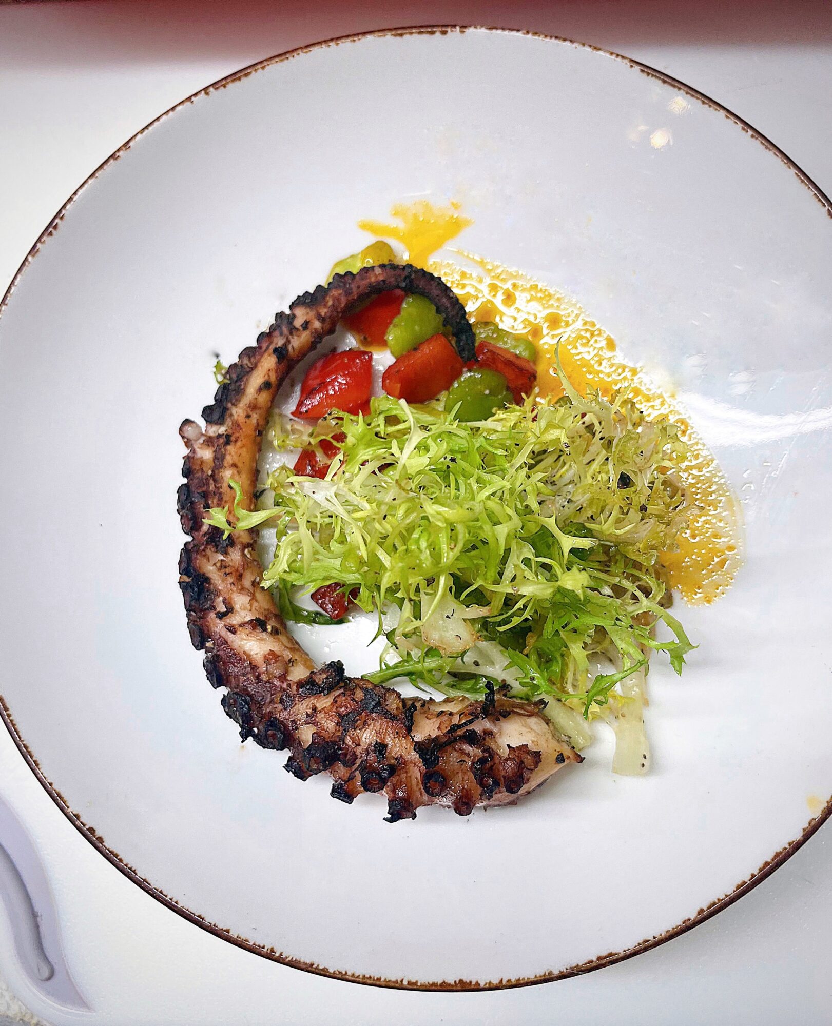 Grilled octopus tentacle with salad, cherry tomatoes, and lemon on a white plate with a gold rim.