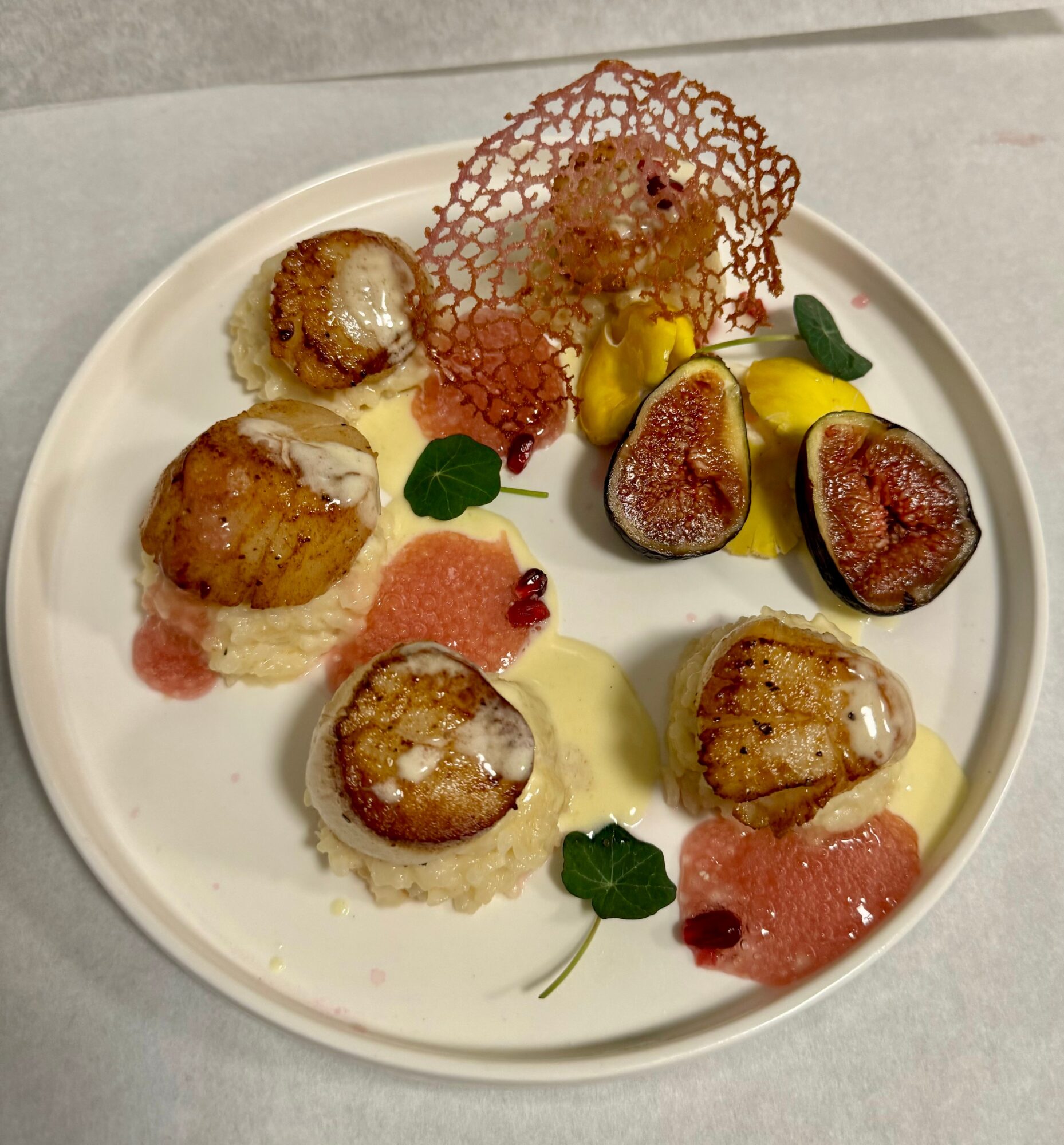 Plate with seared scallops, figs, colorful sauces, and decorative crispy garnish, arranged artistically on a white dish.