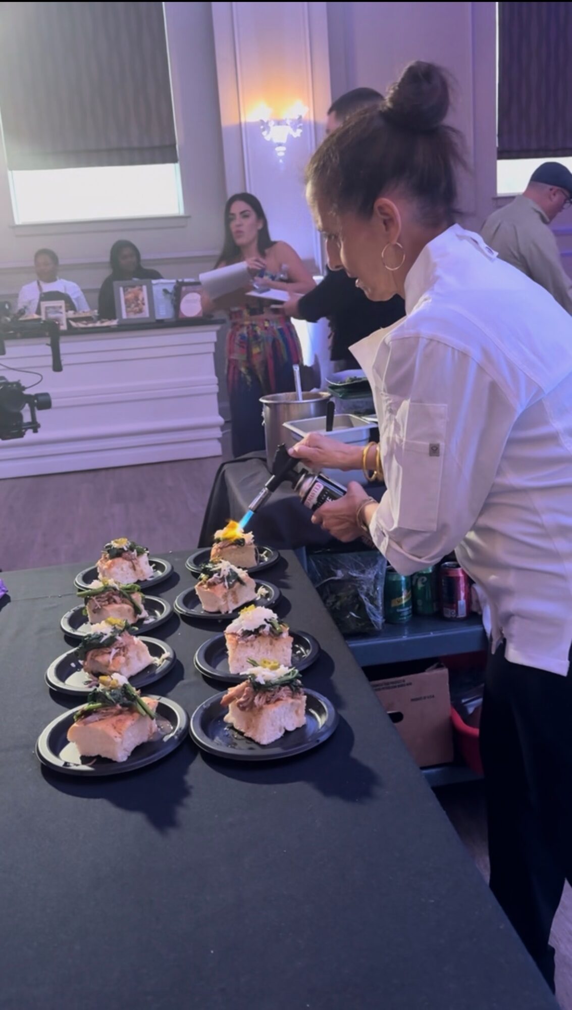 Person preparing plates of food at a buffet table with several dishes, in a room with purple lighting and people in the background.