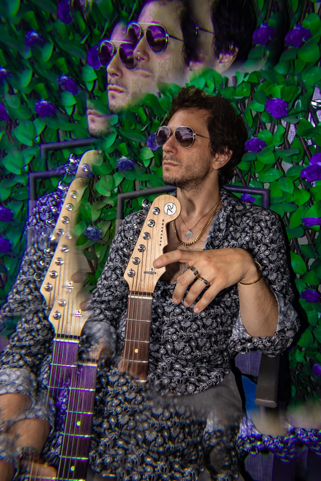 Man with sunglasses and patterned shirt sitting with two guitars, surrounded by green foliage and purple flowers.