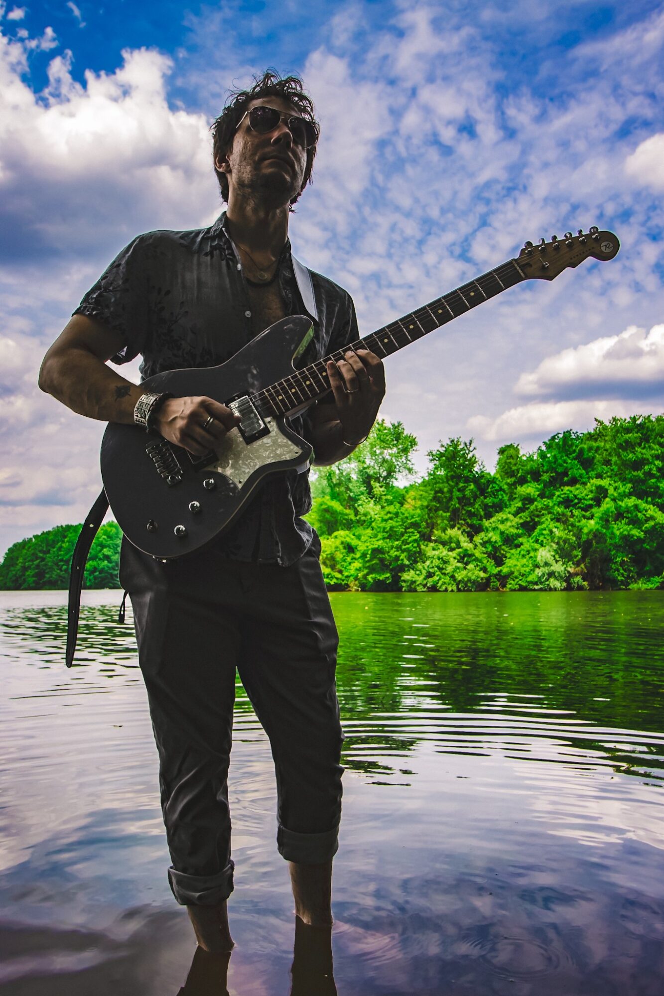 Person standing in water holding an electric guitar outdoors with a sky and trees in the background.