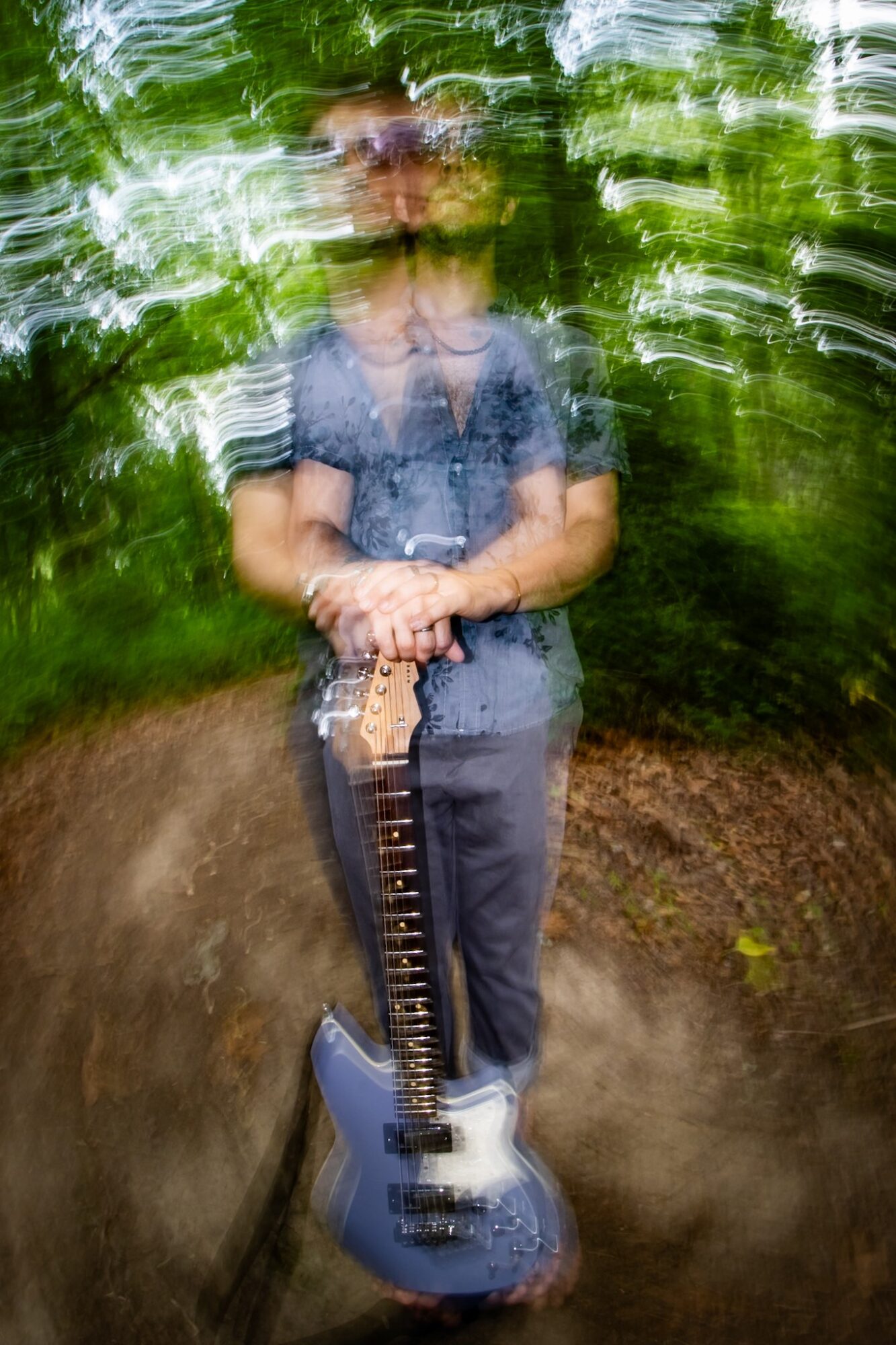 Person holding a guitar outdoors with trees and greenery, motion blur effect, standing on a dirt path.