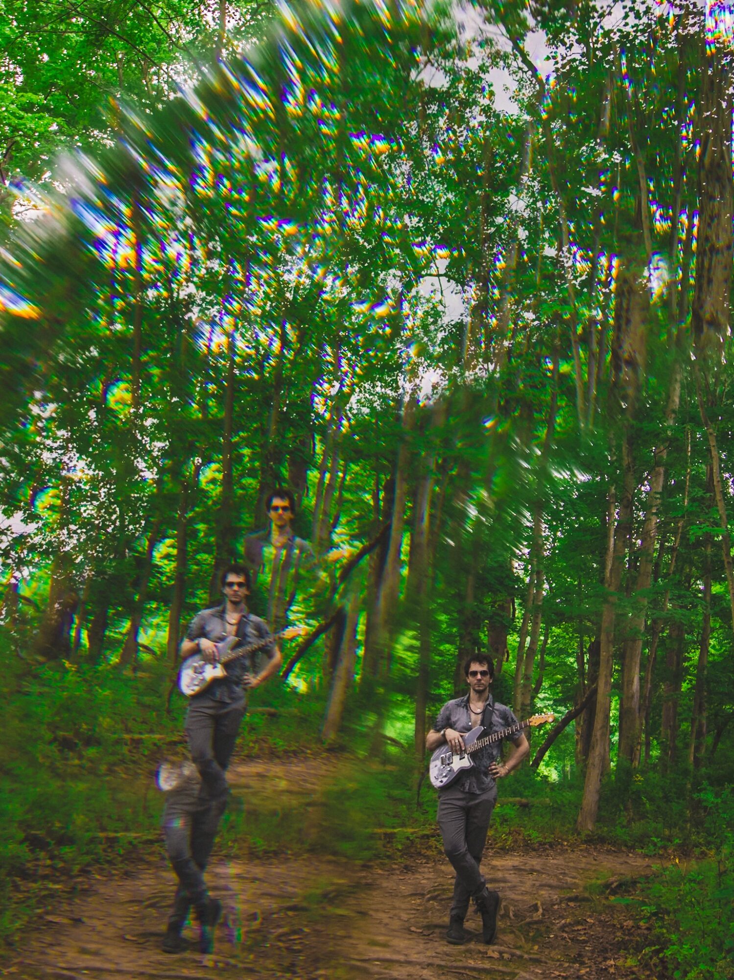 Three people playing guitars in a forest with green trees and a dirt path, sunlight filtering through the leaves.