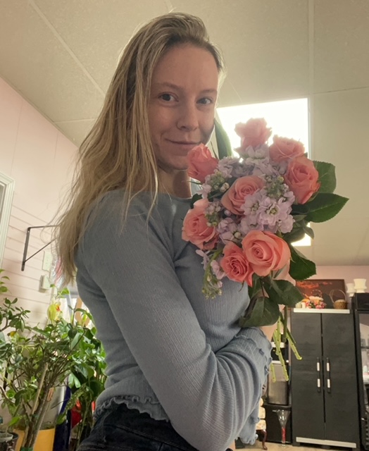 Woman holding a bouquet of pink and purple flowers, smiling, indoors with plants and cabinets in background.