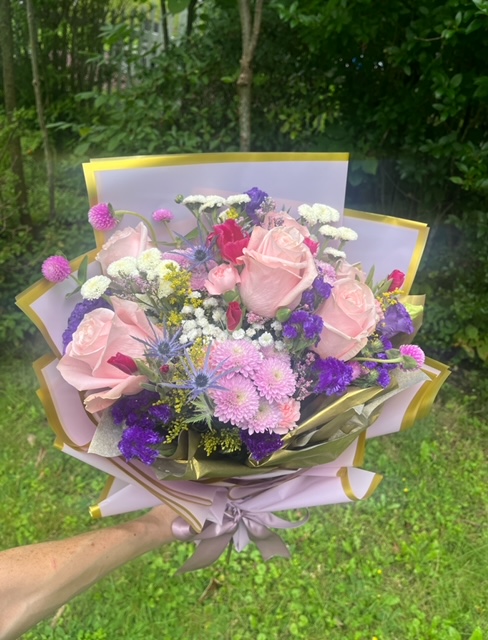 Person holding a colorful bouquet of pink, purple, and white flowers with green leaves, outdoors on grass.