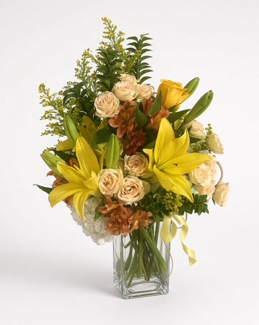 Flower arrangement with yellow lilies, cream roses, and greenery in a clear glass vase.