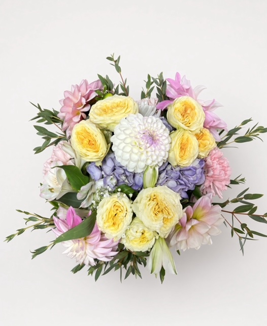 Round bouquet of yellow, pink, purple, and white flowers with green leaves on a white background.