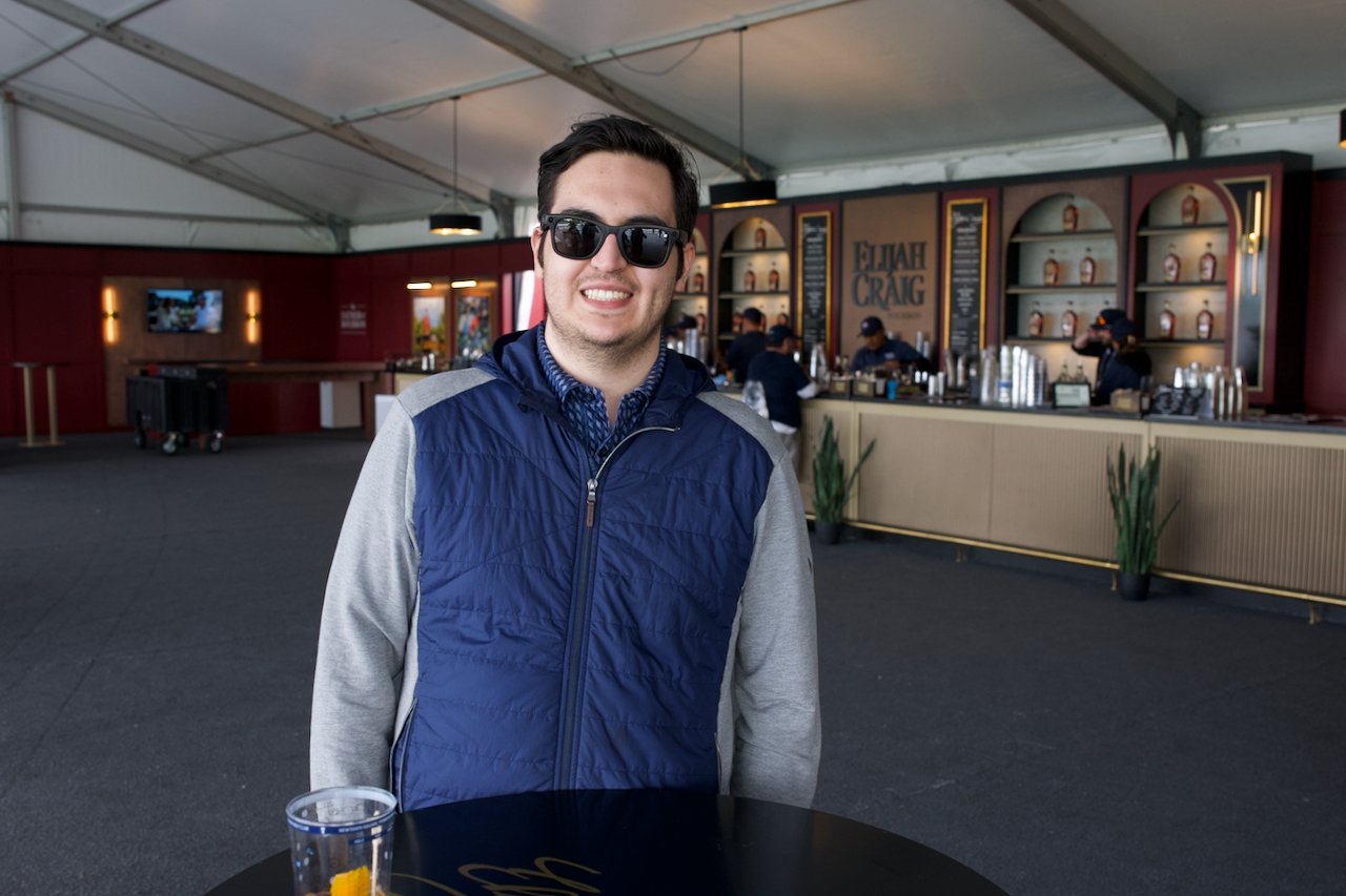 Smiling man wearing sunglasses and a blue vest inside a large event tent with a bar in the background.