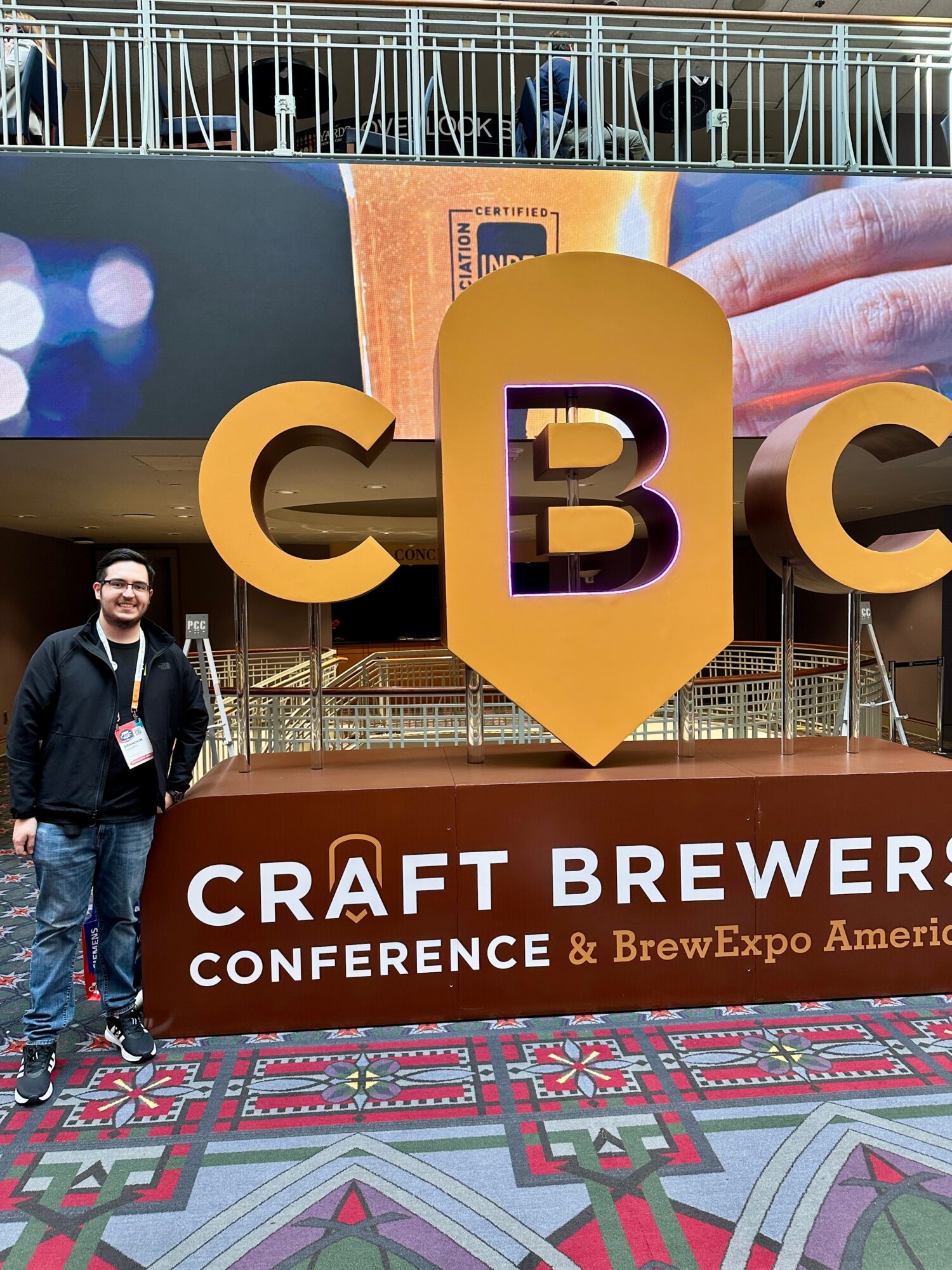Person standing next to large CBC sign at craft brewing conference, with patterned carpet and conference banner in background.
