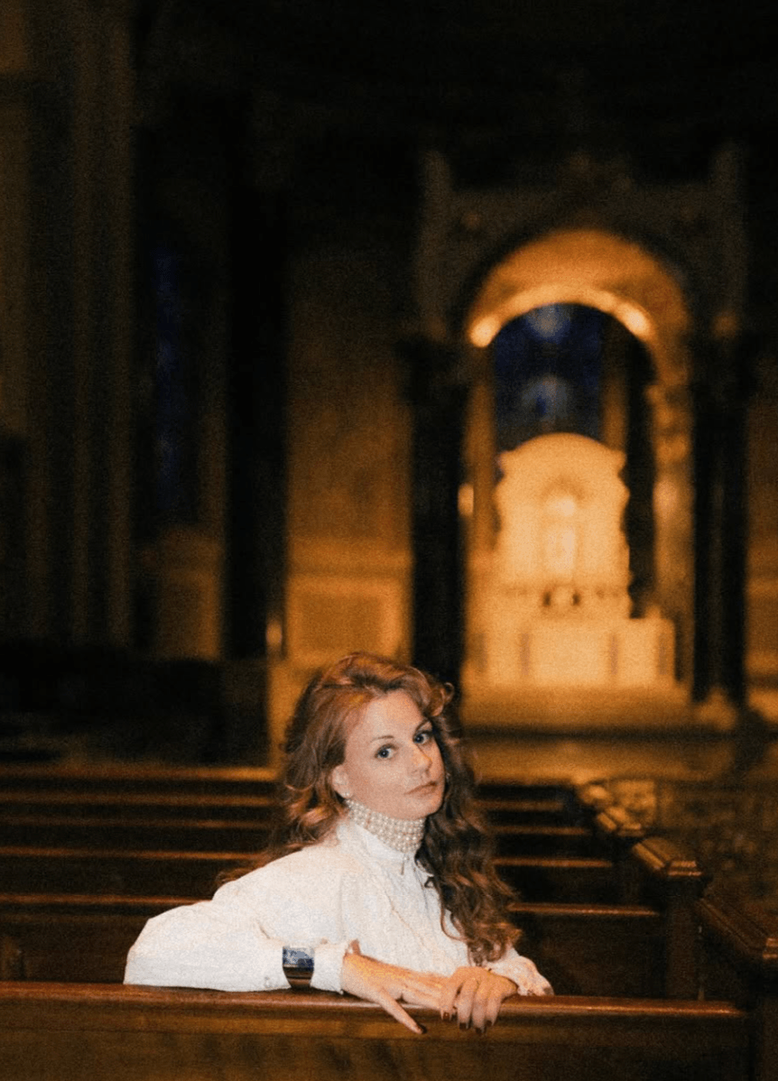 Woman with long hair sitting in a church or cathedral interior with ornate architecture and an altar in the background.