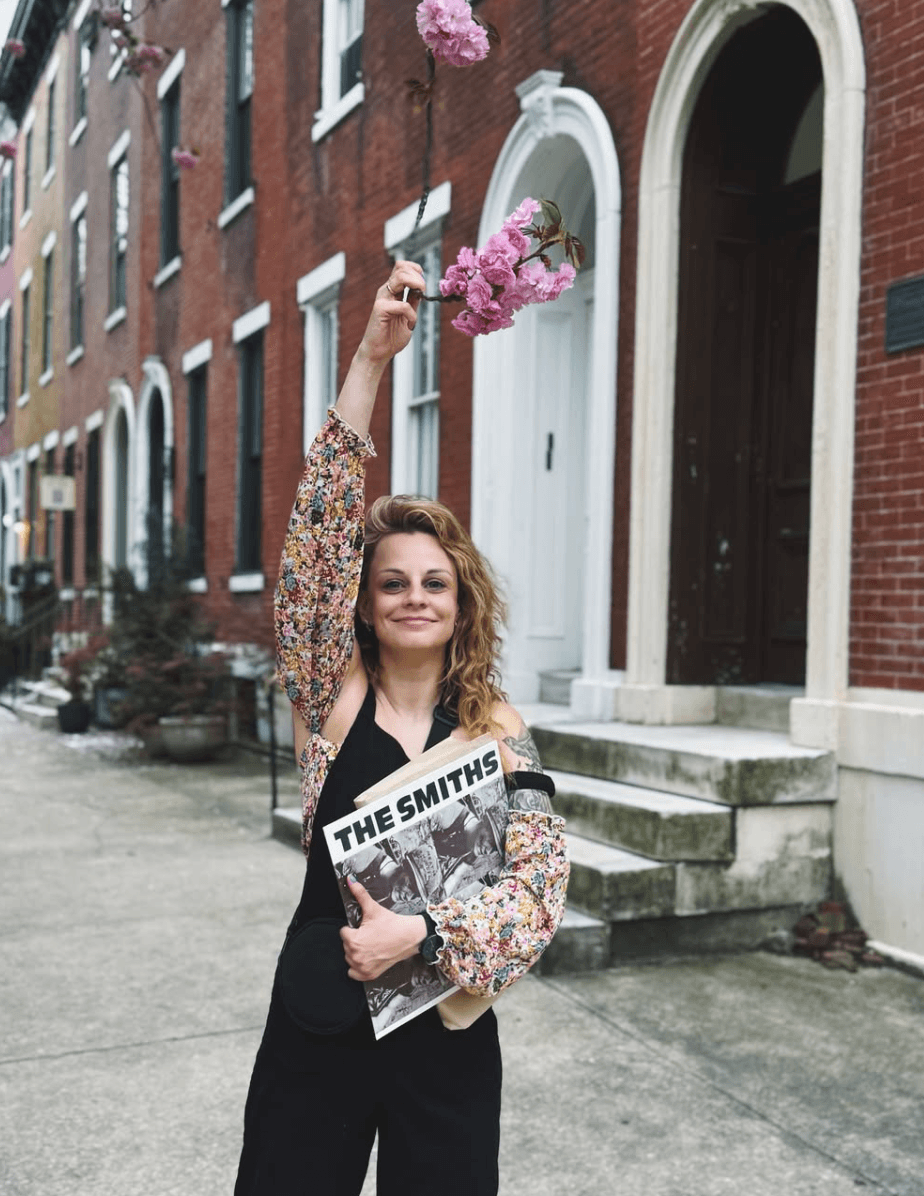 Woman holding a newspaper and a branch of pink flowers on a city sidewalk, smiling, with brick buildings in background.