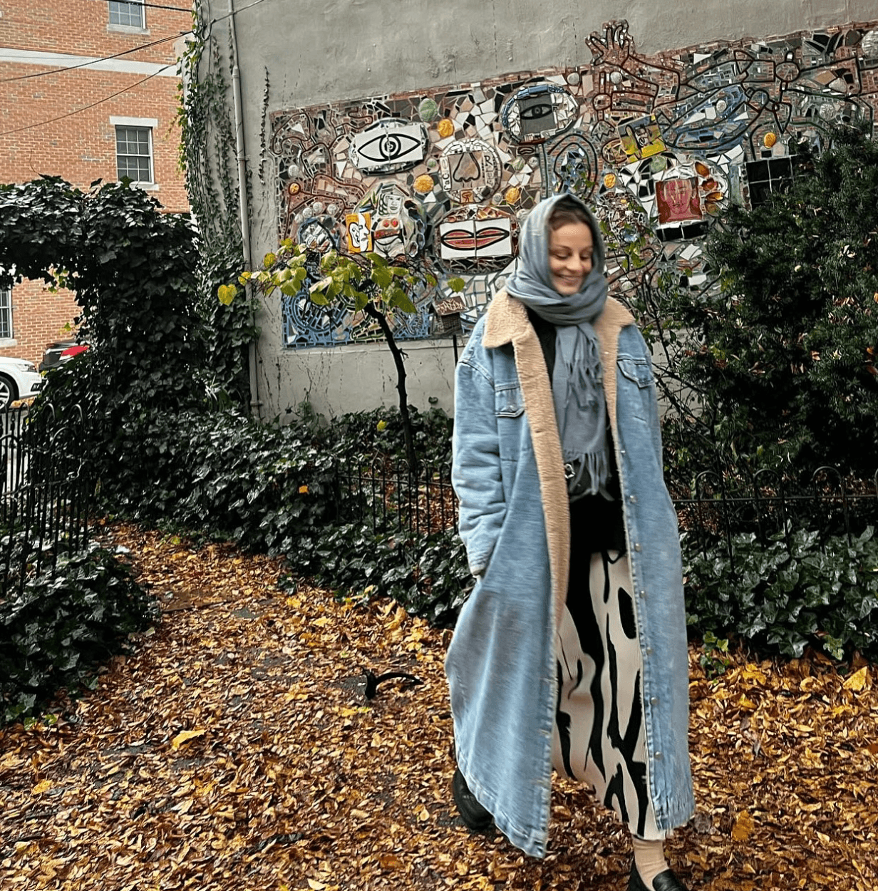 Young woman with a headscarf and long coat standing on a leaf-covered path in front of a colorful mural, surrounded by greenery.