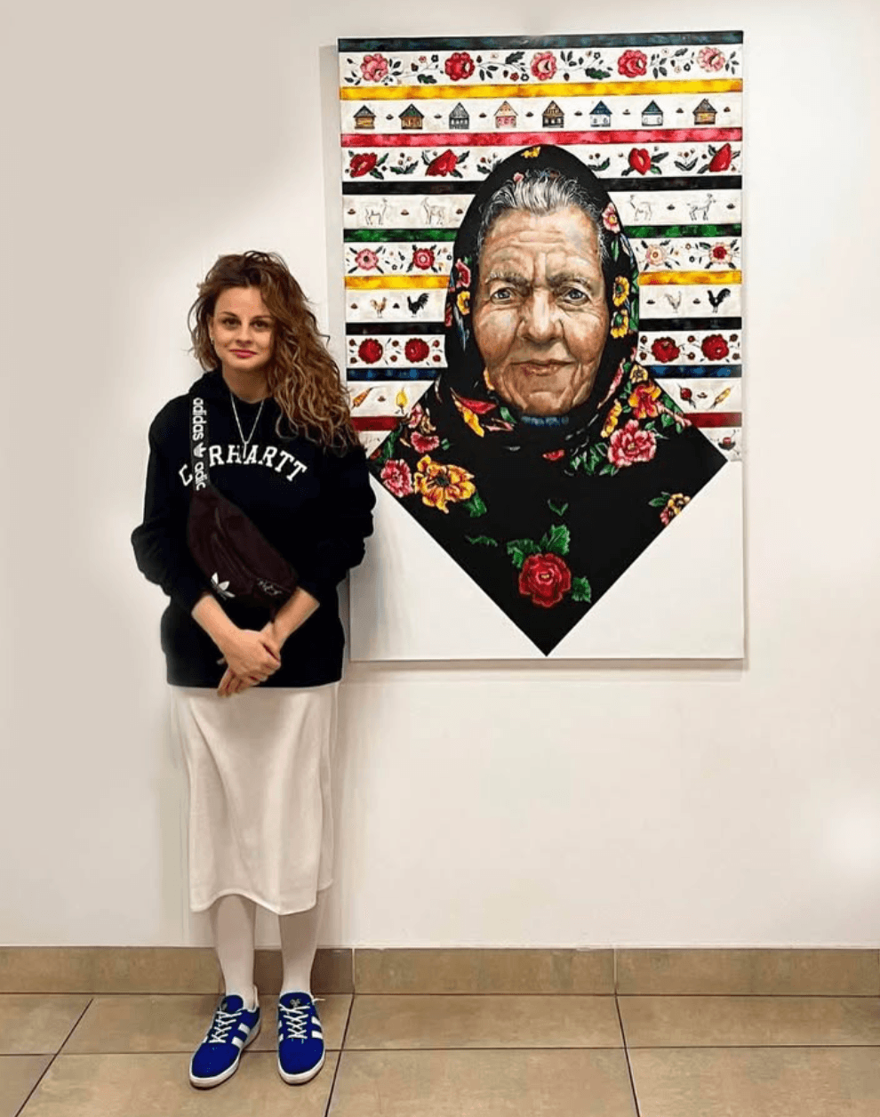 Young woman with curly hair standing next to a colorful portrait of an elderly woman wearing a floral scarf, on a white wall.