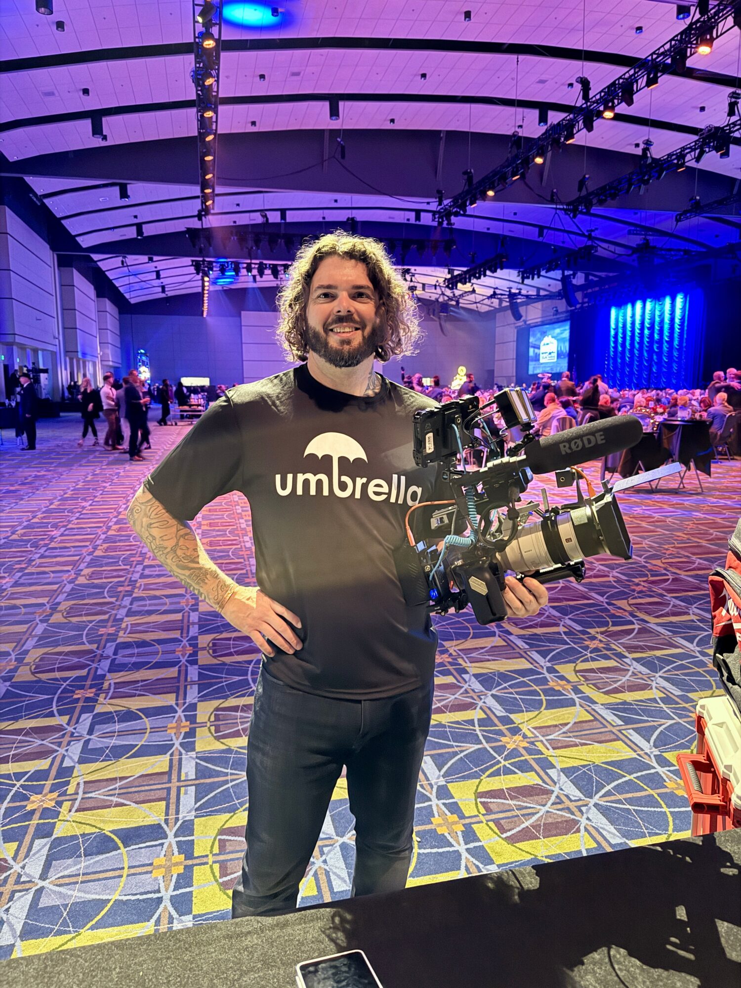 Man with curly hair and beard holding a professional video camera in a large event hall with colorful lighting.