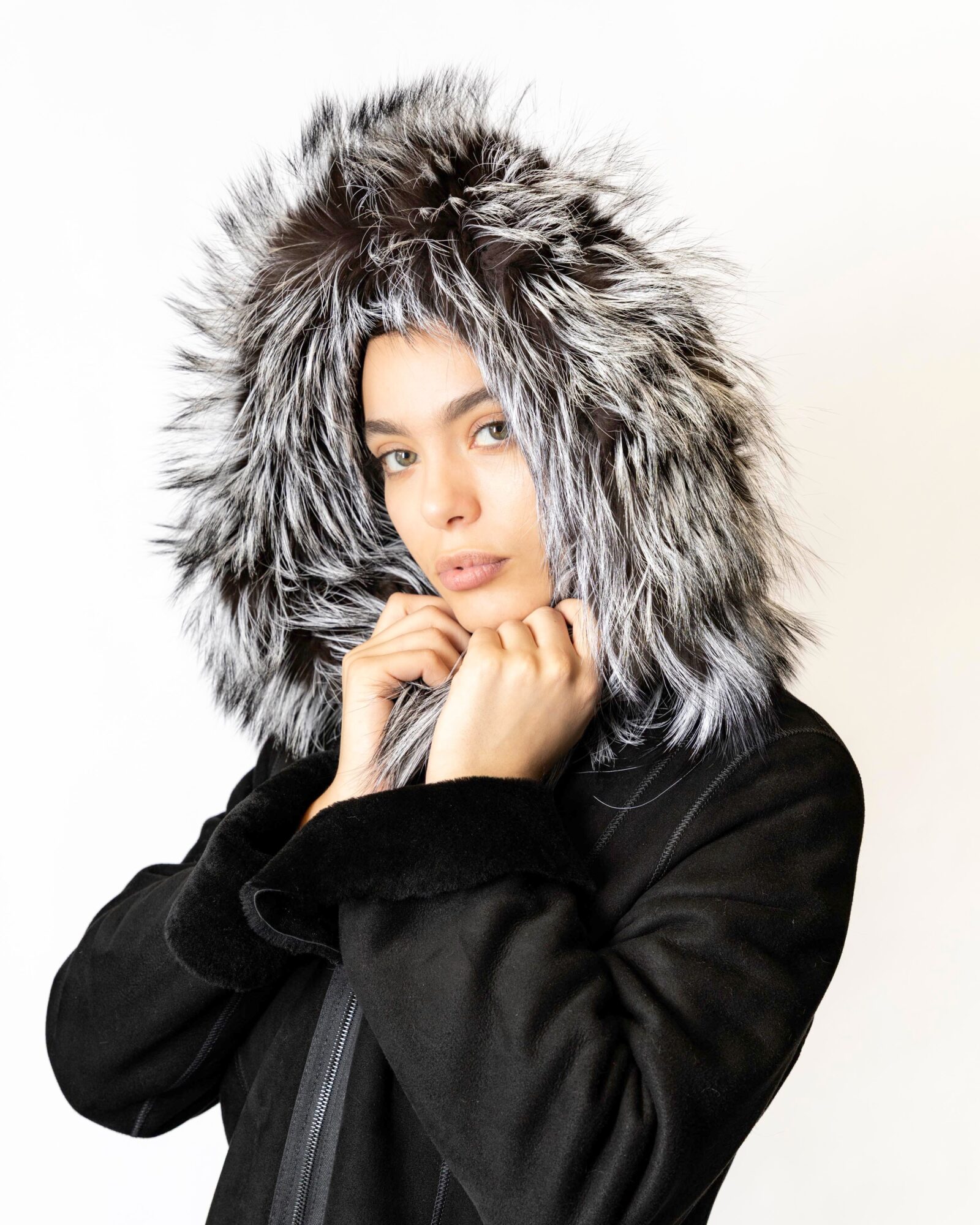 Woman wearing a black coat with a large fur-lined hood, looking at the camera, white background.