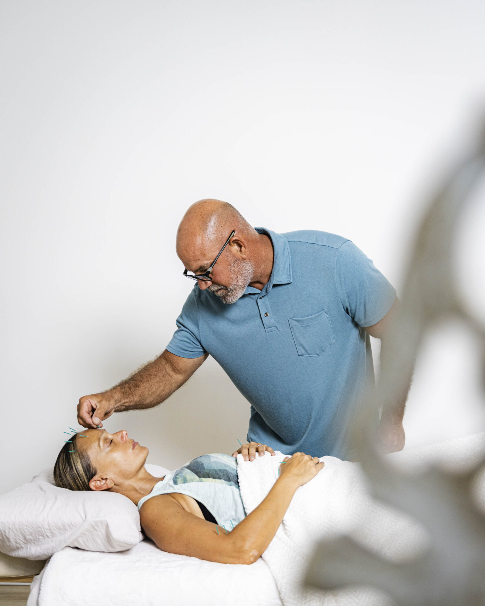 A man in a blue shirt leans over a woman lying in bed, smiling and looking at each other.