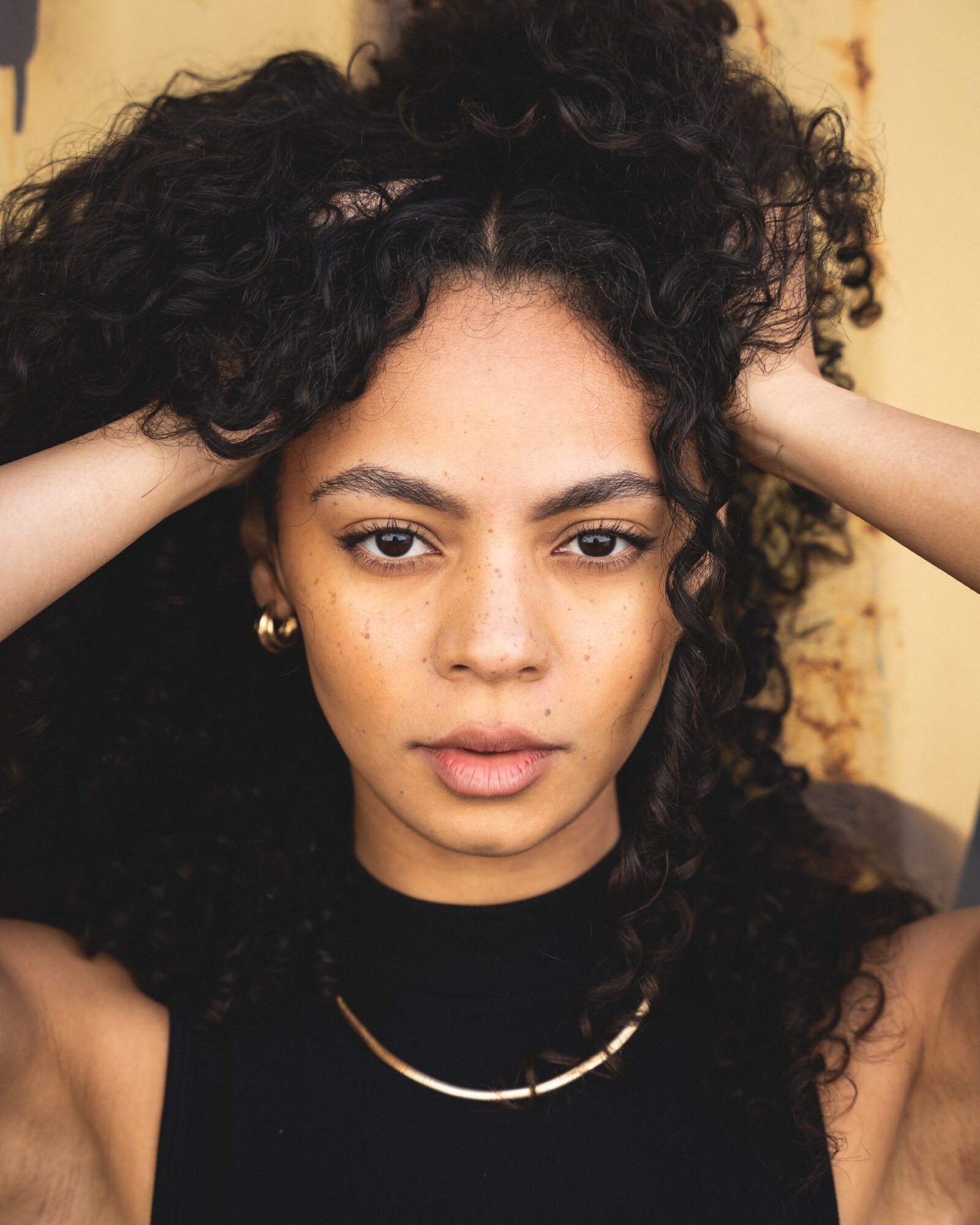 Young woman with curly hair holding her head, wearing a black top and jewelry, looking directly at the camera.