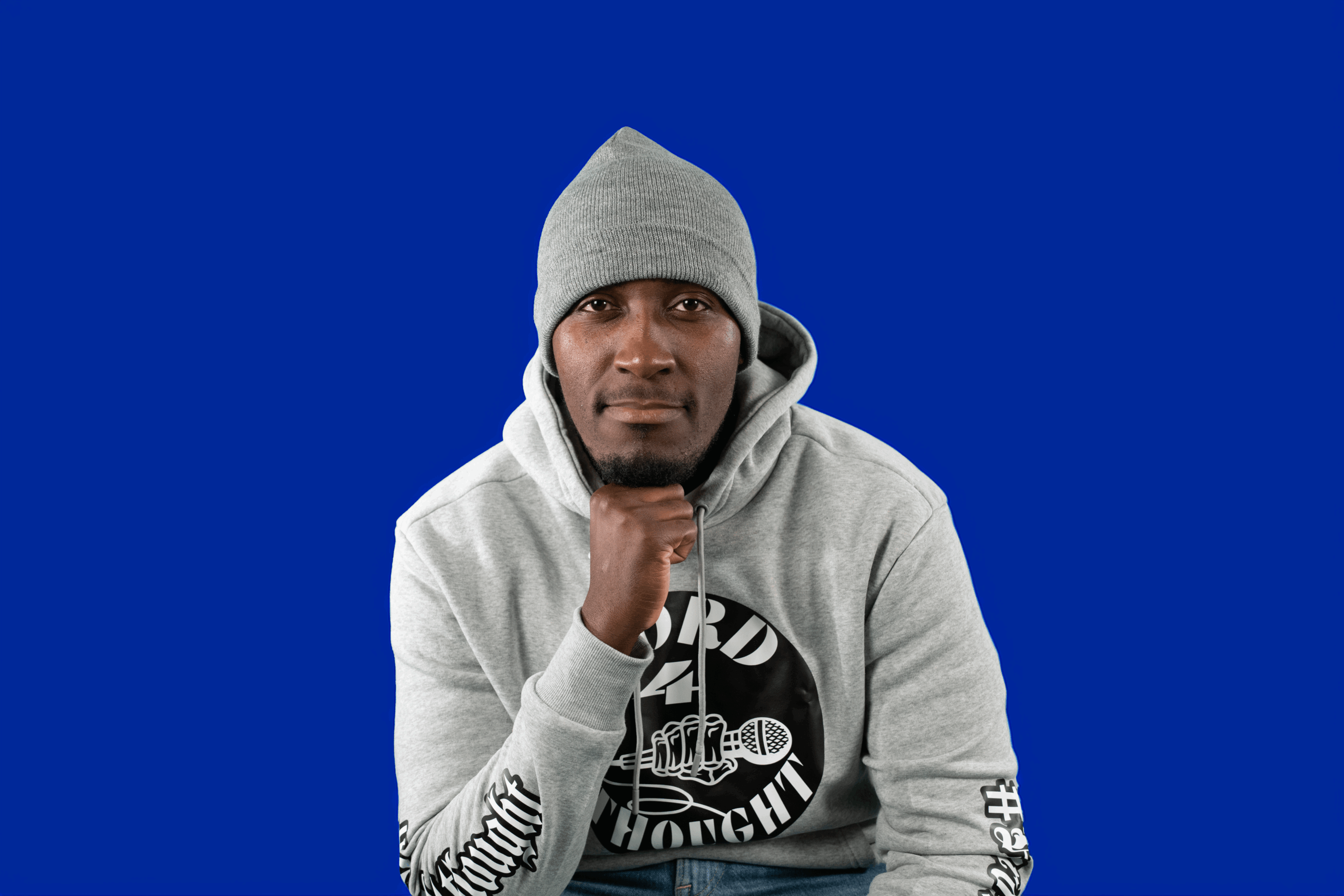 Young man wearing a gray hoodie and beanie, standing against a blue background, looking at the camera with a neutral expression.