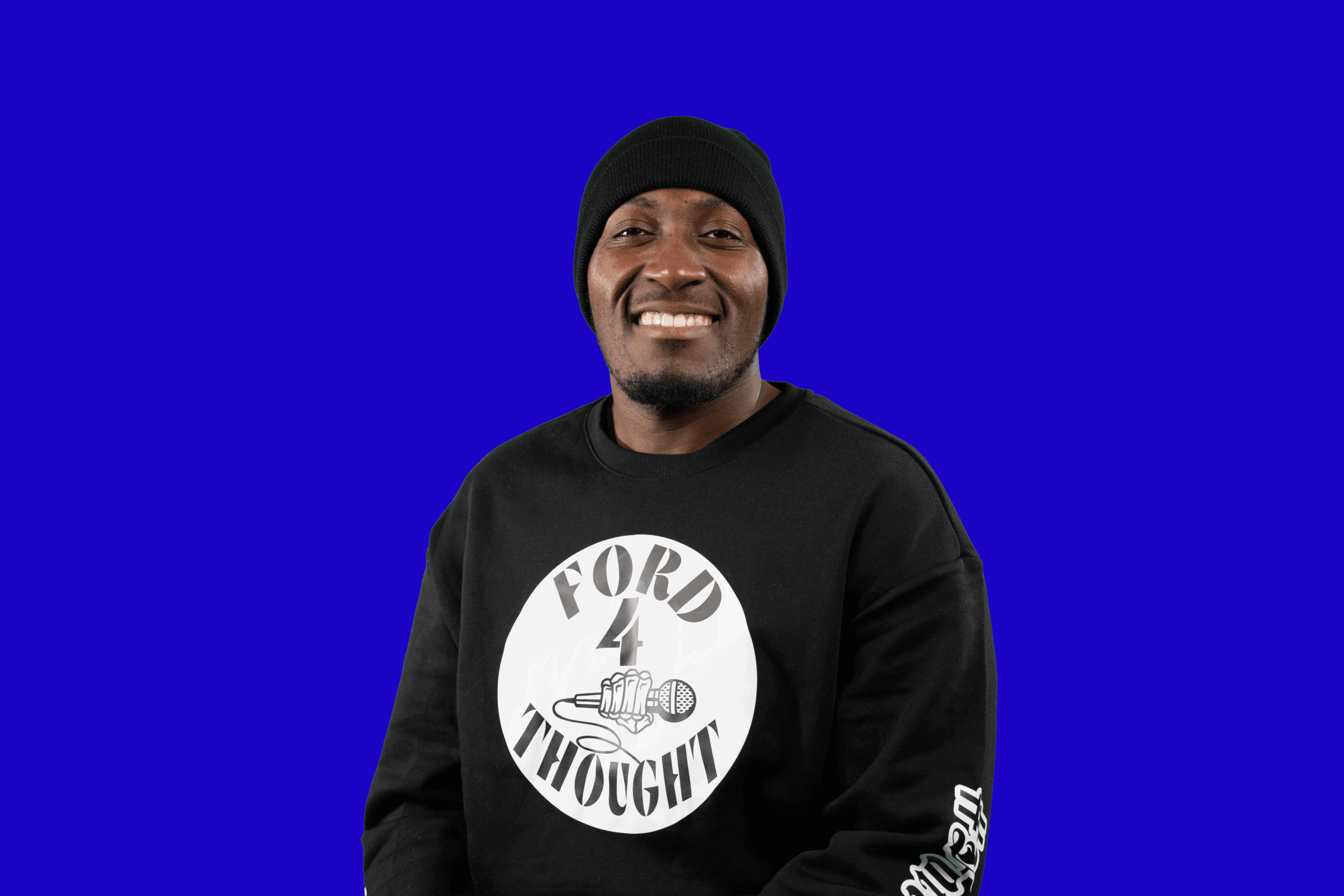 Smiling man wearing a black beanie and sweatshirt with a white circular logo, standing against a blue background.
