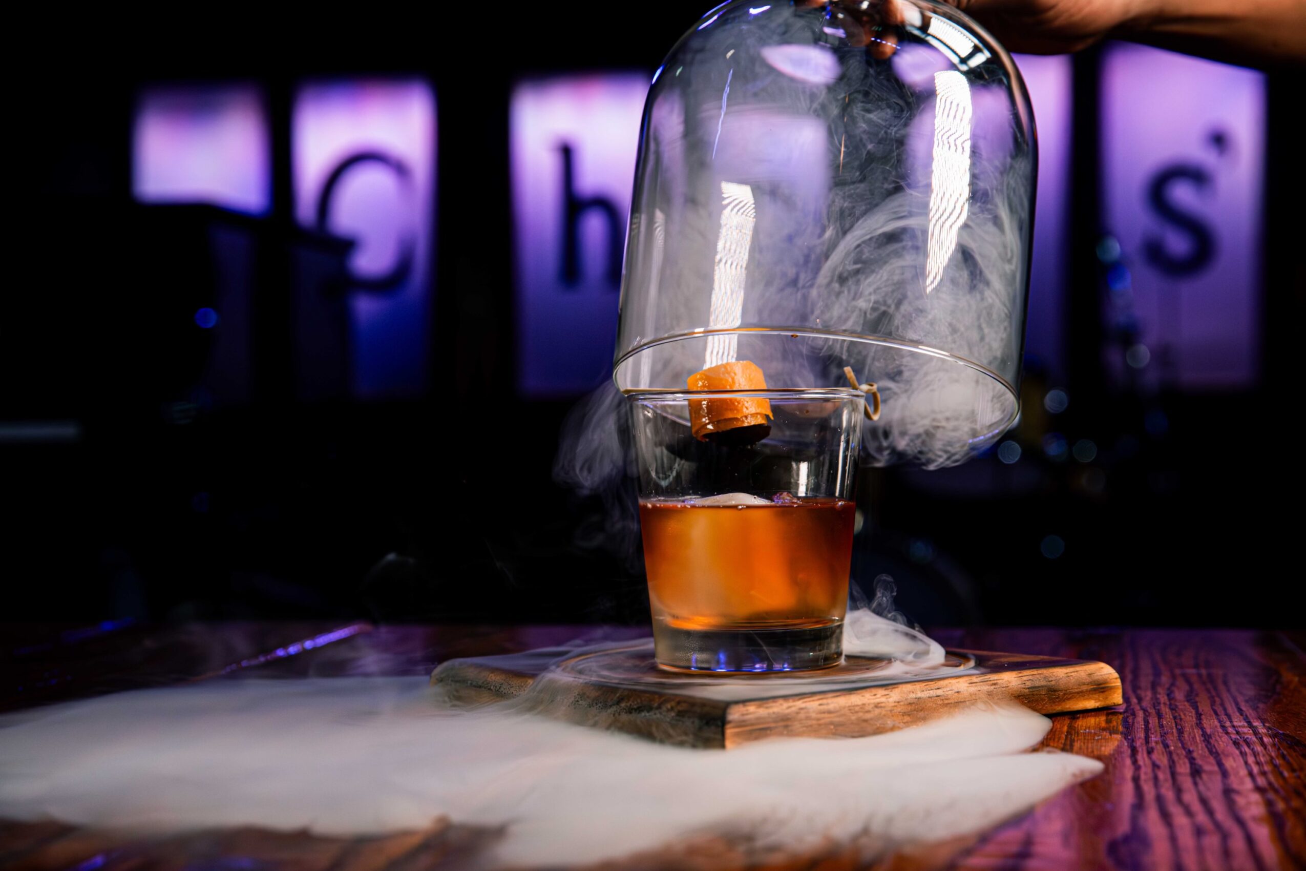 A glass dome covers a small glass with orange liquid, placed on a wooden board on a table, with a blurred background.