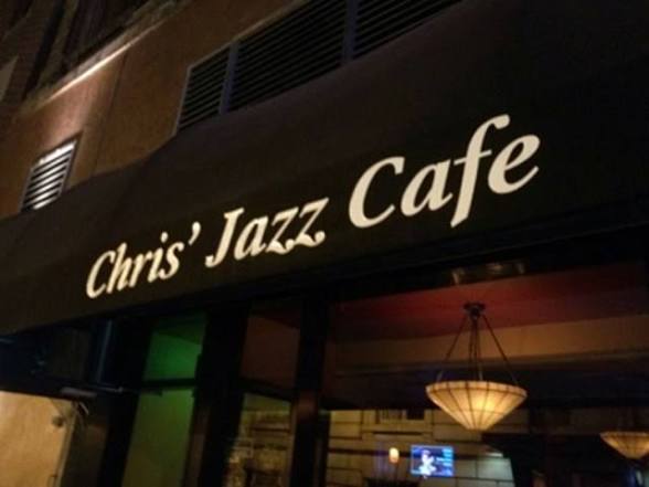 Sign for Chris' Jazz Cafe with black background and white text, interior visible through window, chandelier hanging inside.