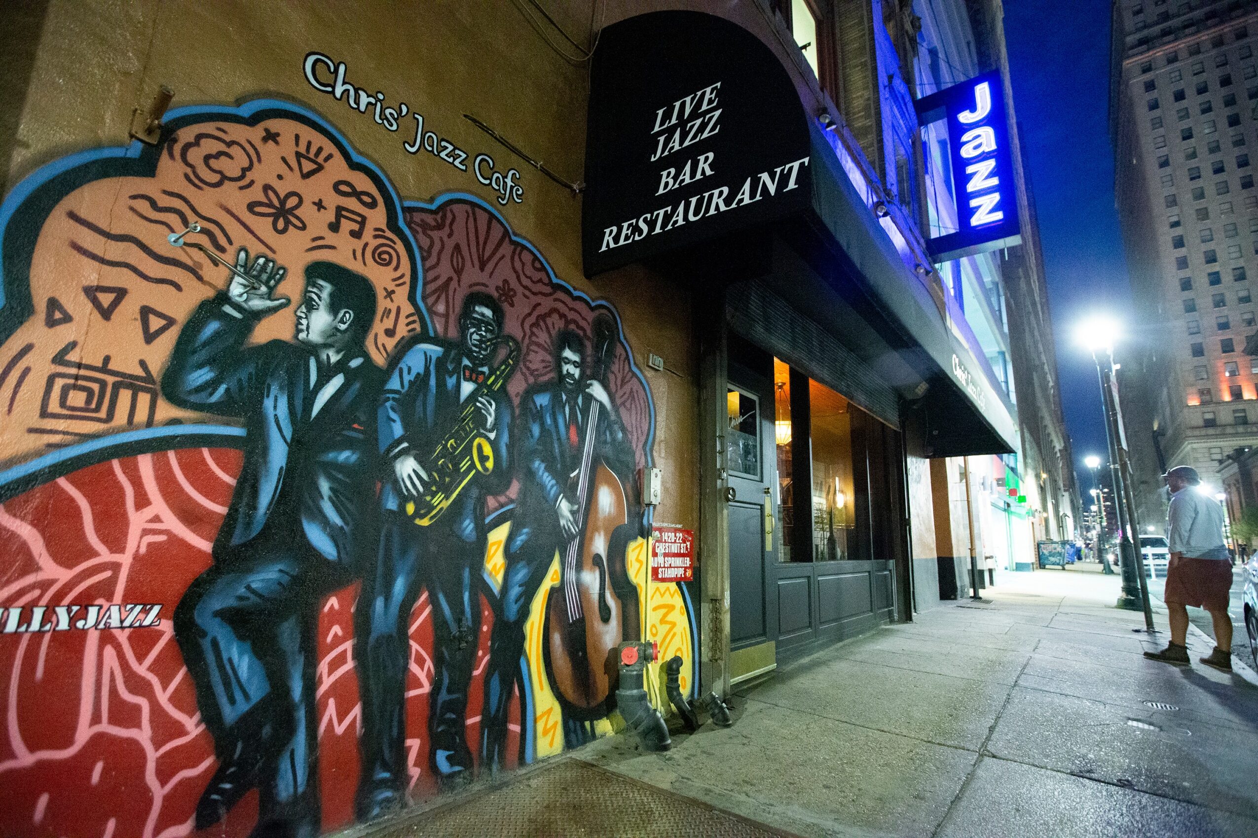 Street view of a jazz bar with a colorful mural of musicians playing instruments, illuminated signs, and pedestrians.
