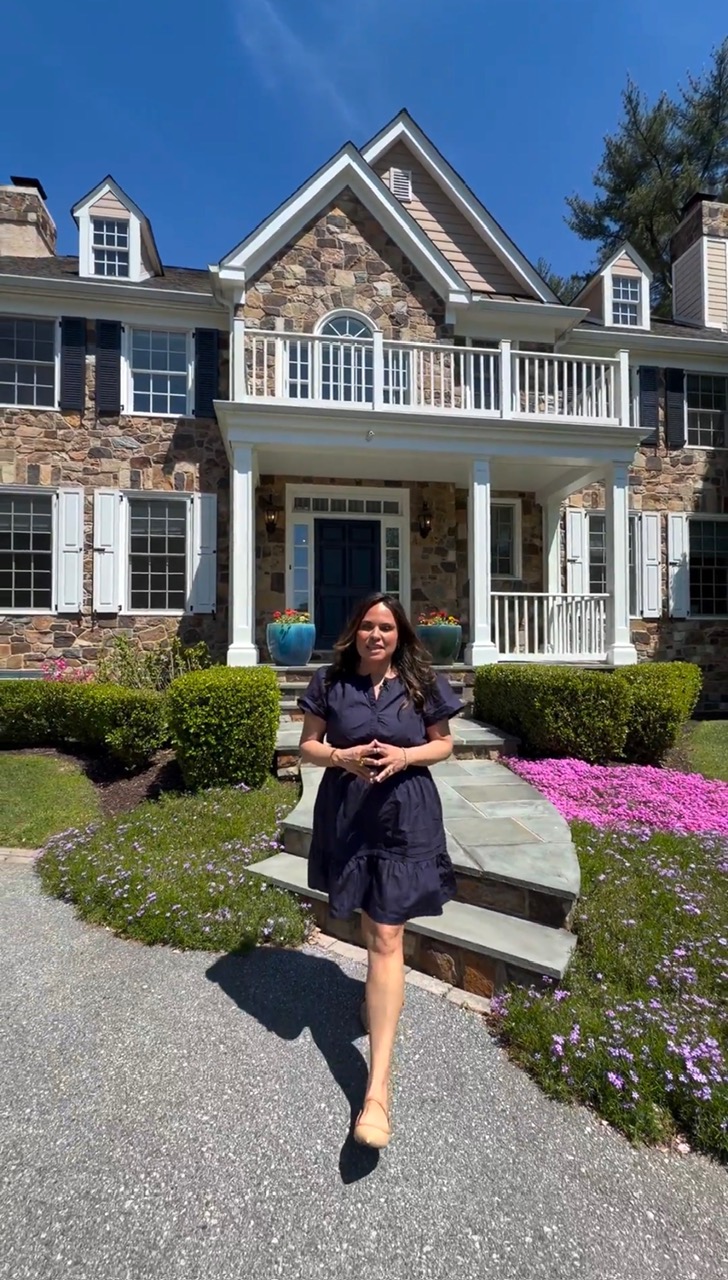 Woman in dark dress standing on a stone pathway in front of a large brick house with white trim and a balcony, under a blue sky.