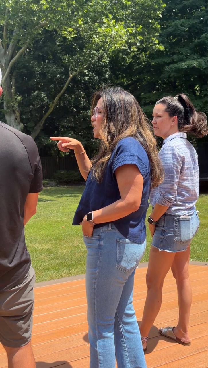 Three women standing outdoors on a grassy area with trees, engaged in conversation, one pointing, all casually dressed.