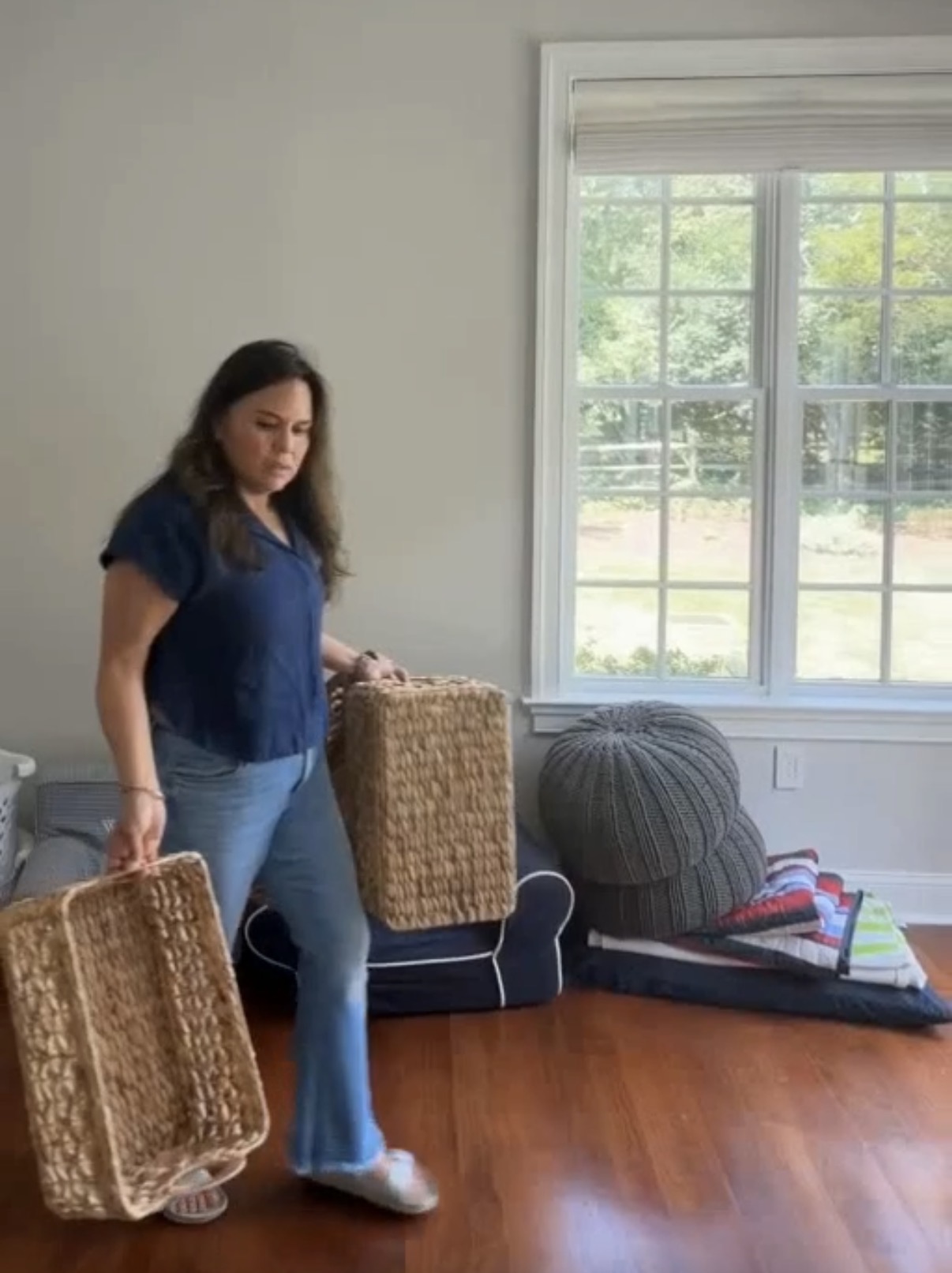 Woman standing in a room holding two woven baskets, with a window and furniture in the background.