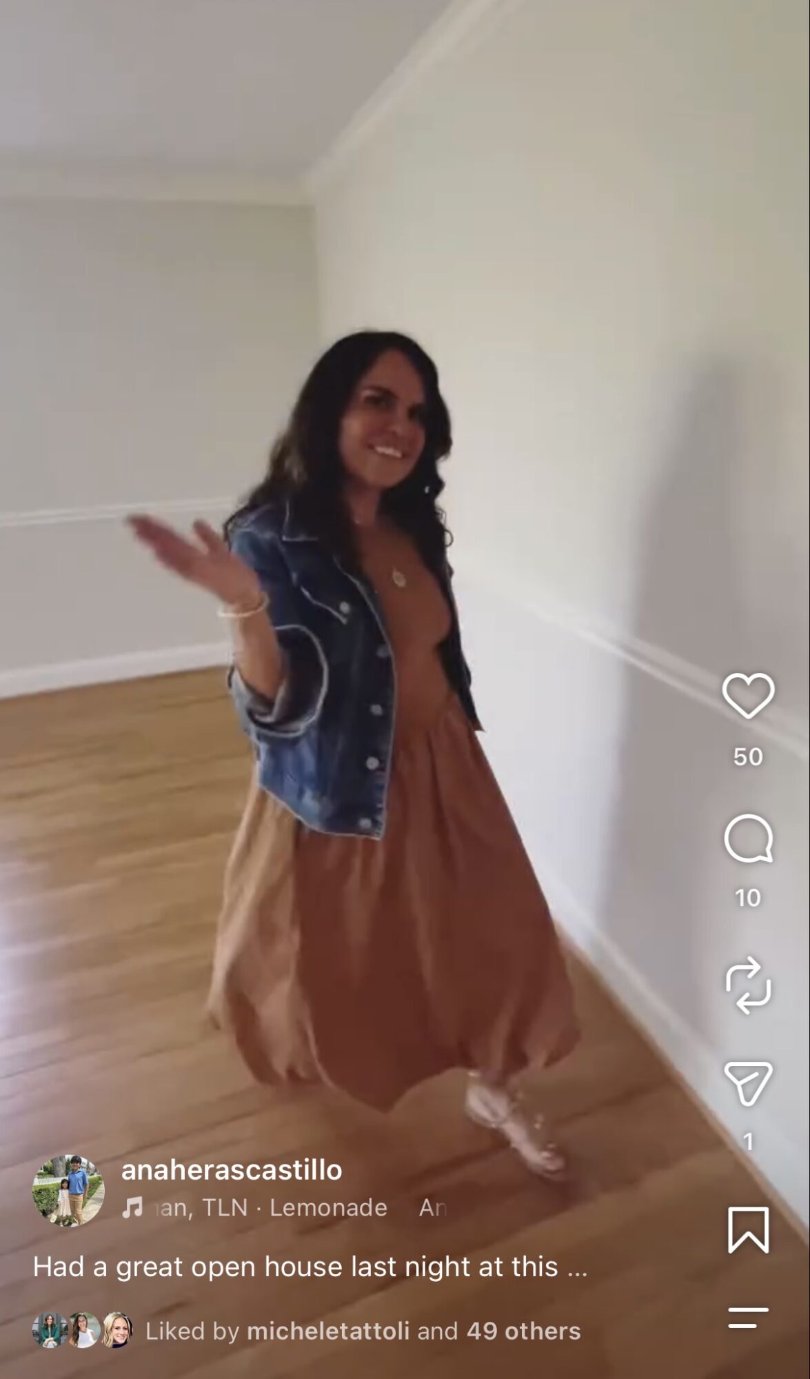 Woman smiling and waving indoors, wearing a denim jacket and brown dress, standing against a white wall.