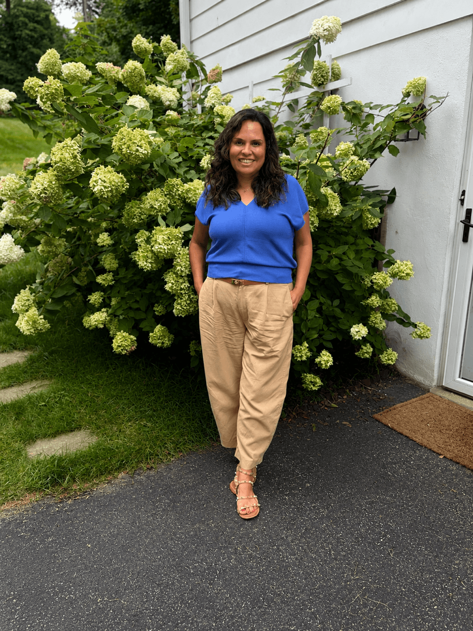 Woman with dark hair in a blue top and beige pants standing outdoors near green bushes and a white house wall.