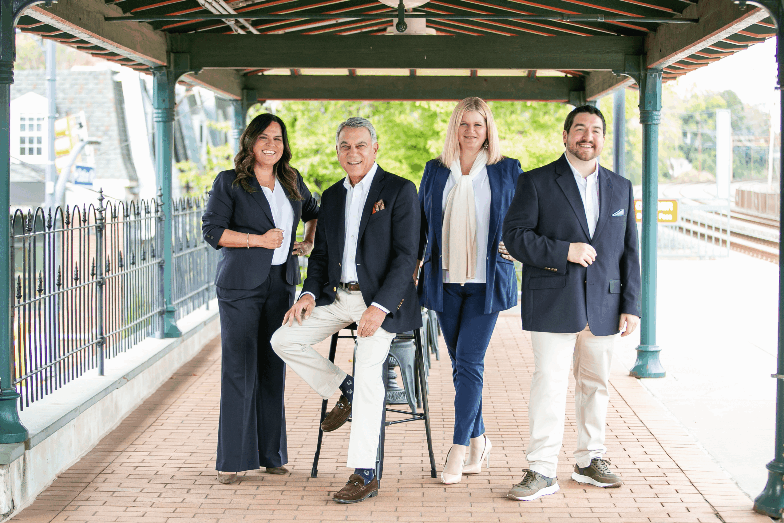 Four people standing under a covered walkway, smiling, with a railing and train tracks in the background.