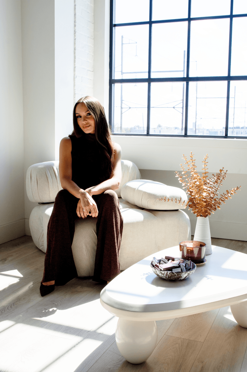 Woman sitting on a white sofa near a large window with sunlight, next to a white coffee table with a vase and snacks.
