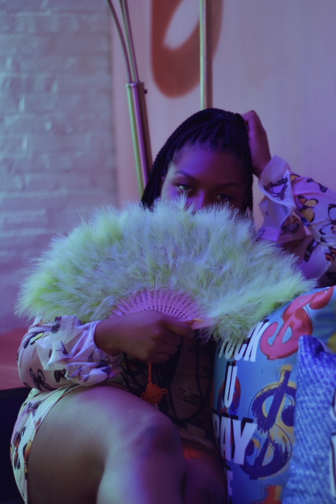 Person holding a green feather fan partially covering their face, sitting indoors with colorful lighting and a bag nearby.